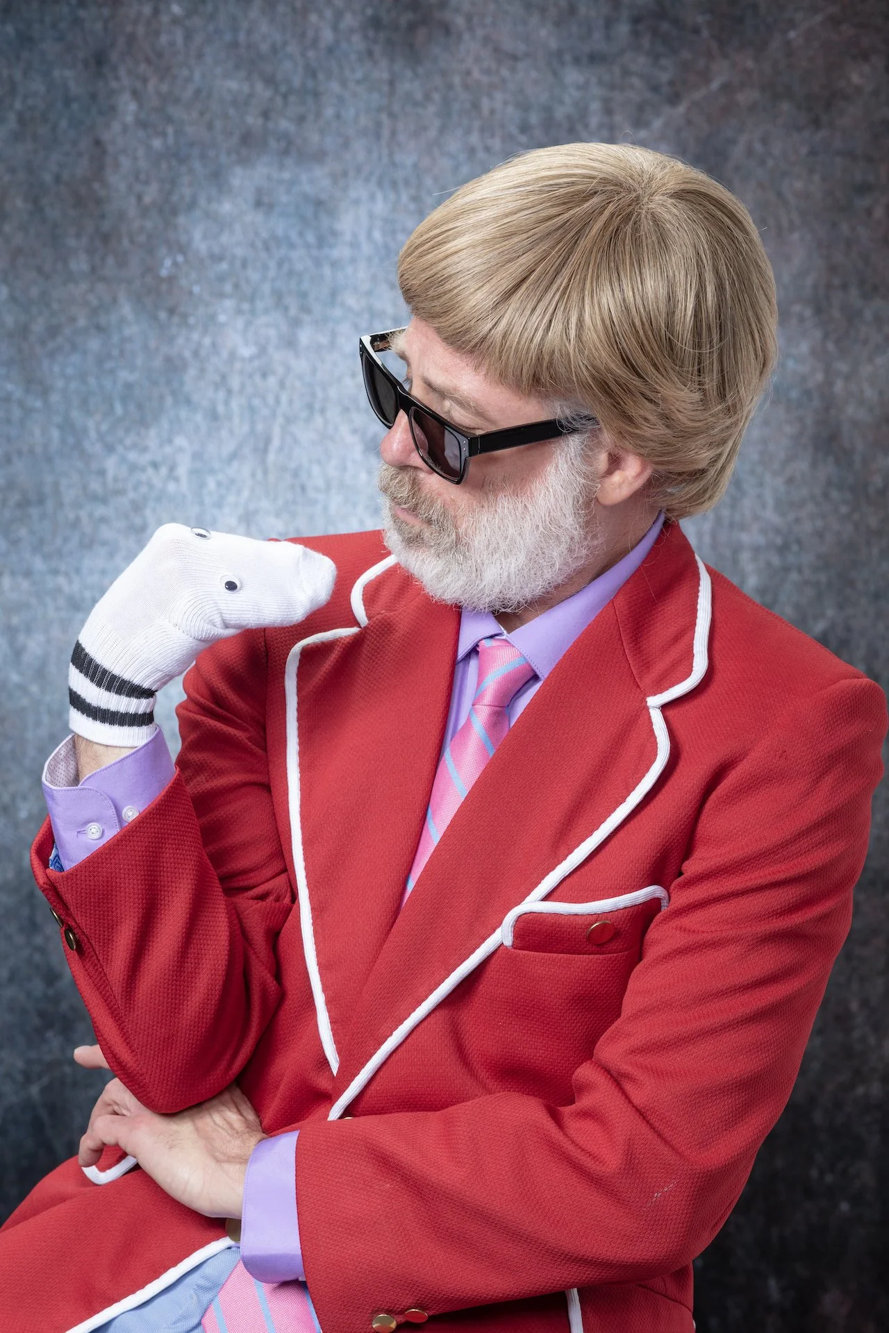 A man with a gray beard and blond hair styled in a mop-top haircut, wearing black sunglasses, a red blazer with white piping, a lavender shirt, and a pink tie with light blue stripes, posing with his left arm crossed and his right arm bent, touching 