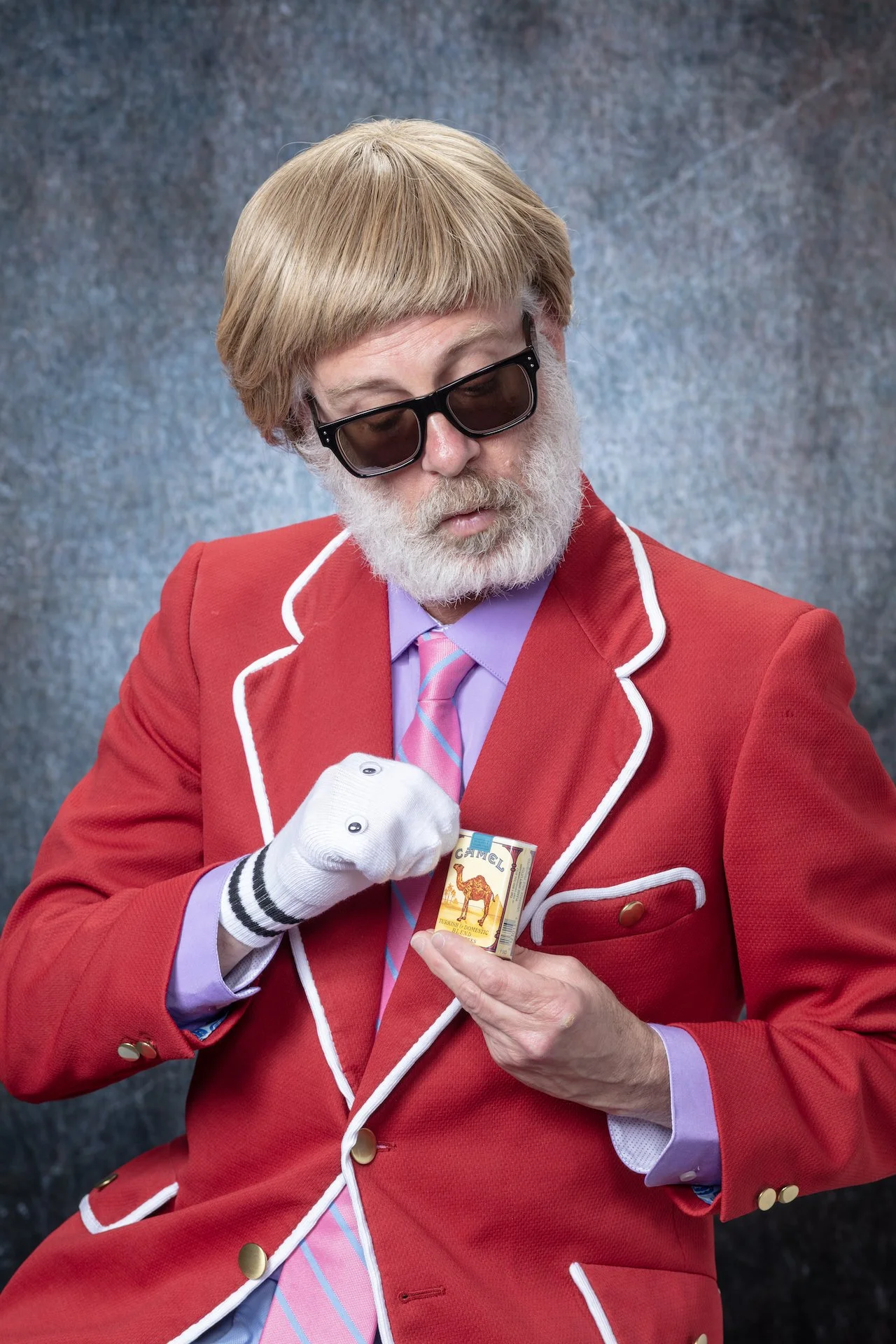 A man with a white beard and light brown hair wearing sunglasses, a red blazer with white trim, a lavender shirt, and a pink striped tie, holding a box of Camel cigarettes.