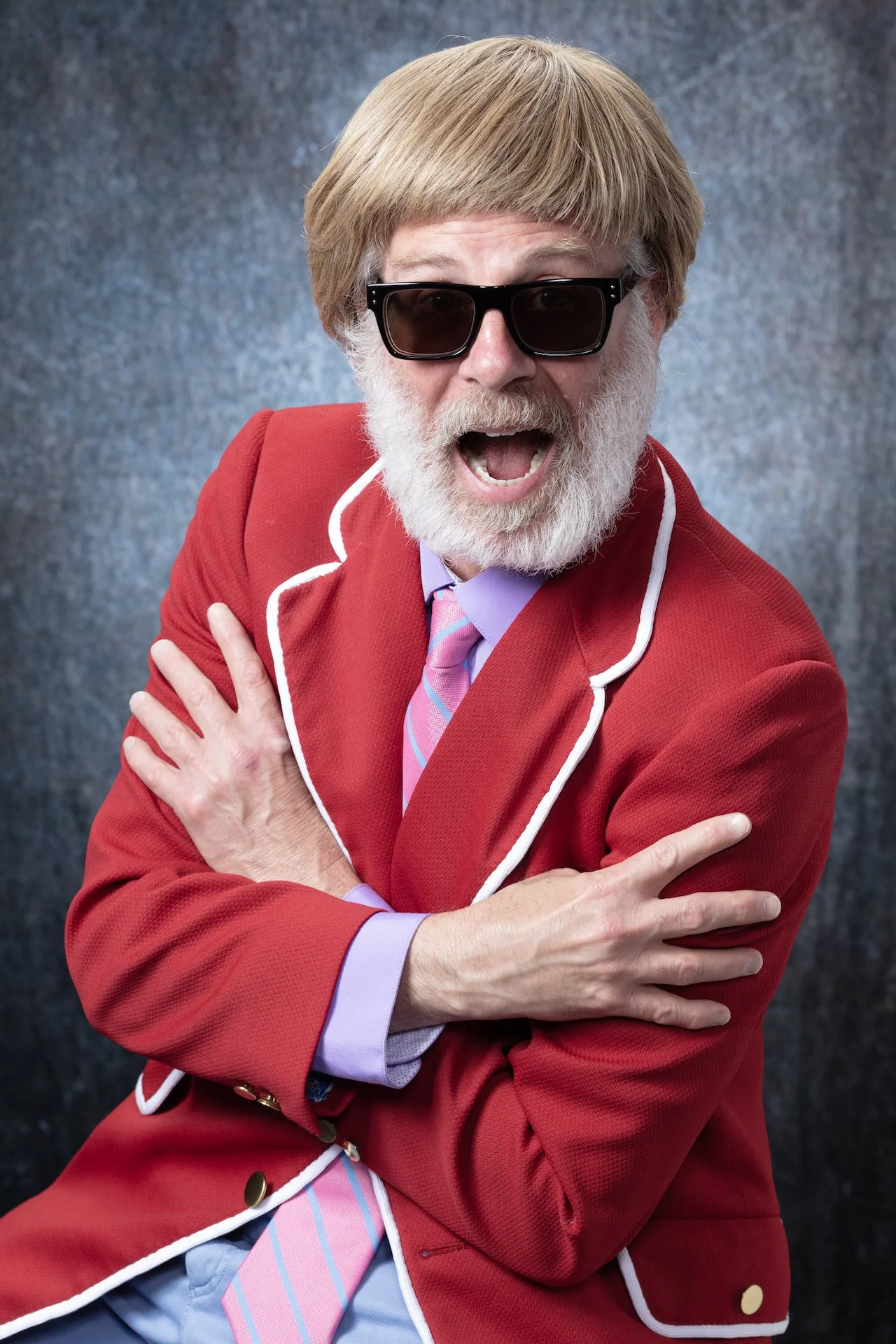A man with a gray beard and hair, wearing black sunglasses, a pink and blue striped tie, a lavender shirt, and a red jacket with white piping, standing against a dark gray background, with his arms crossed.