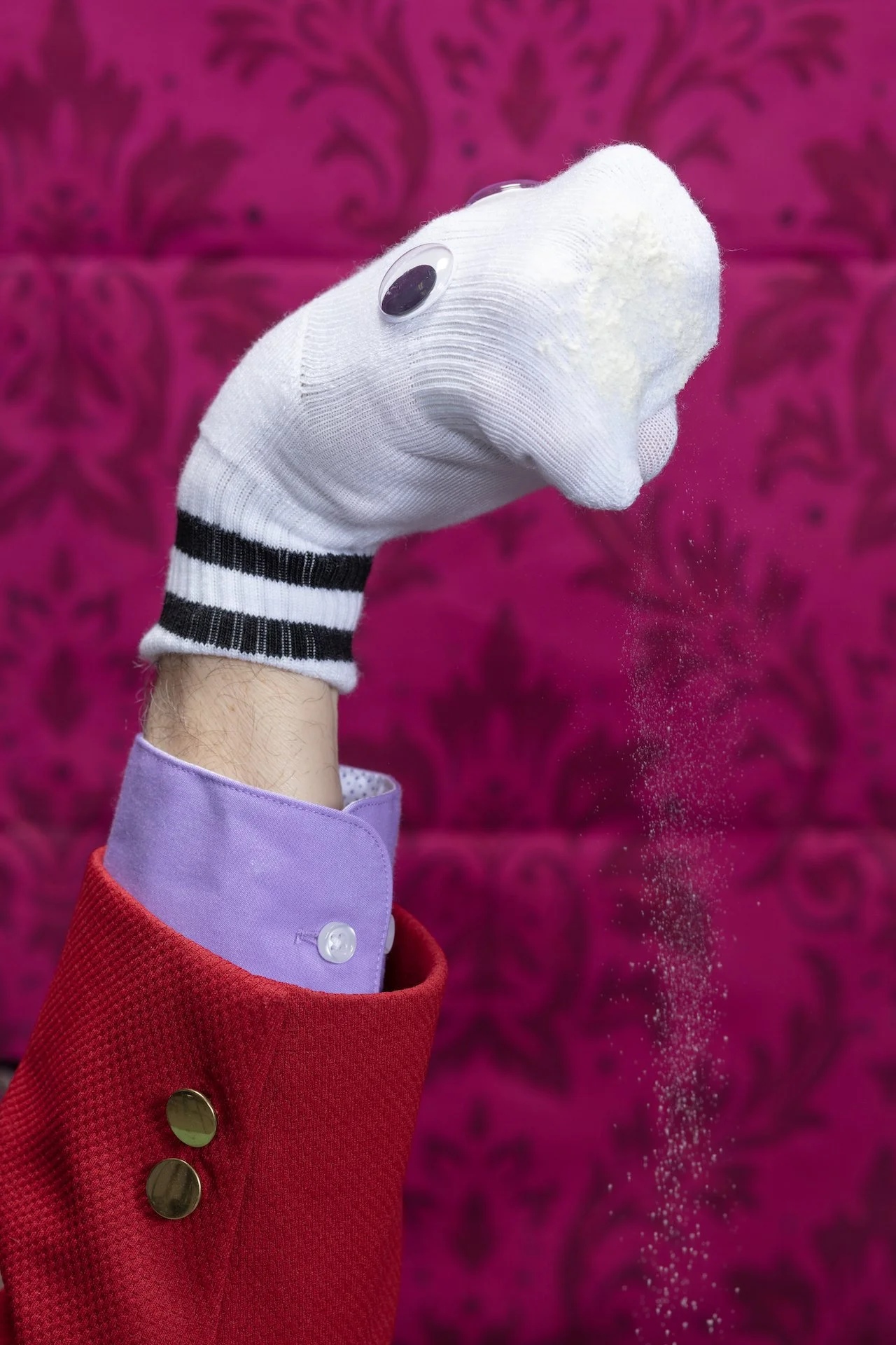 Close-up of a person's hand wearing a white sock glove with googly eye decoration, throwing white powder or glitter against a vibrant pink and purple floral patterned background.