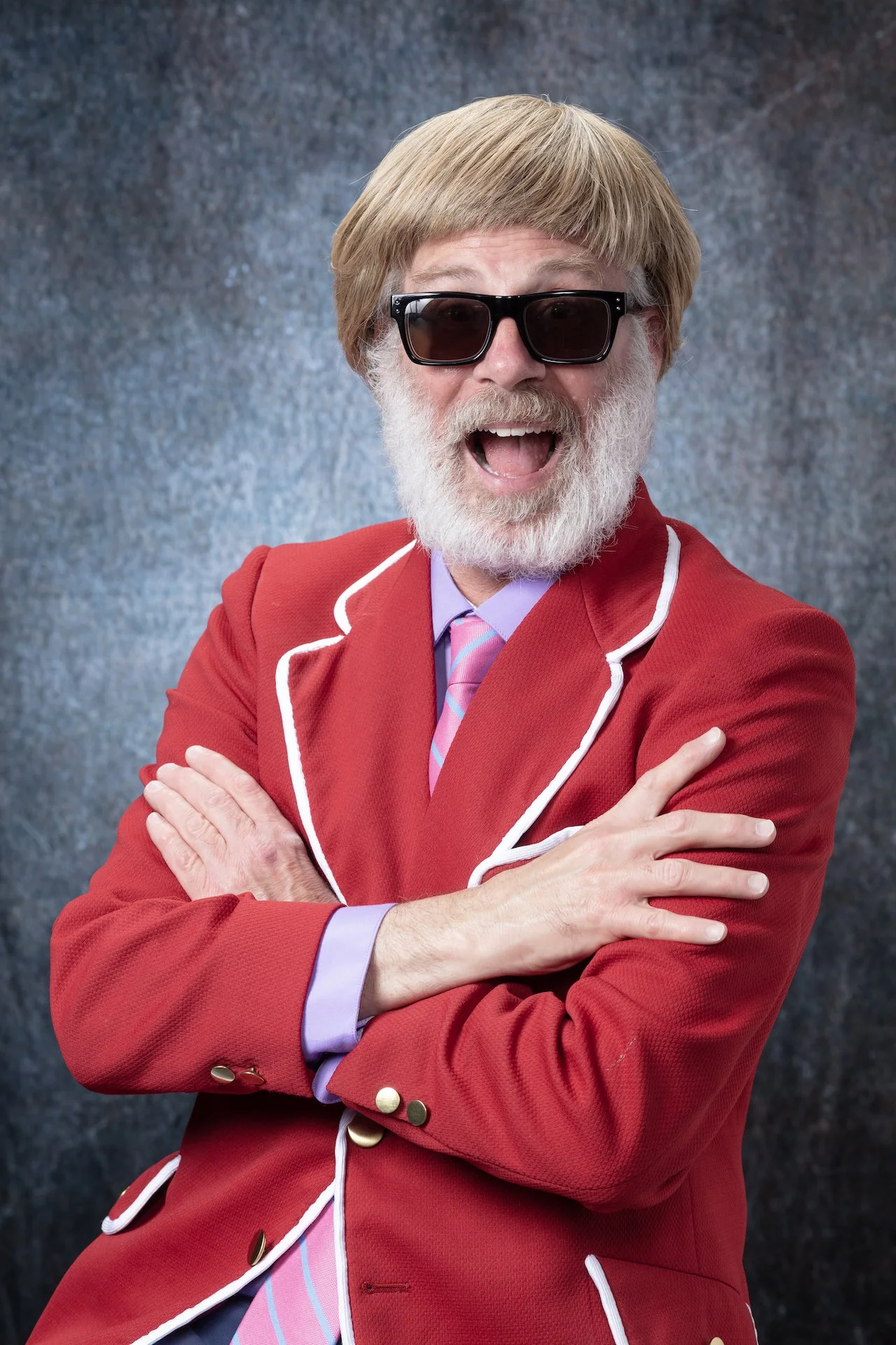 A cheerful man with a white beard, wearing dark sunglasses, a red blazer with white trim, a purple shirt, and a pink striped tie, smiling with arms crossed against a textured background.