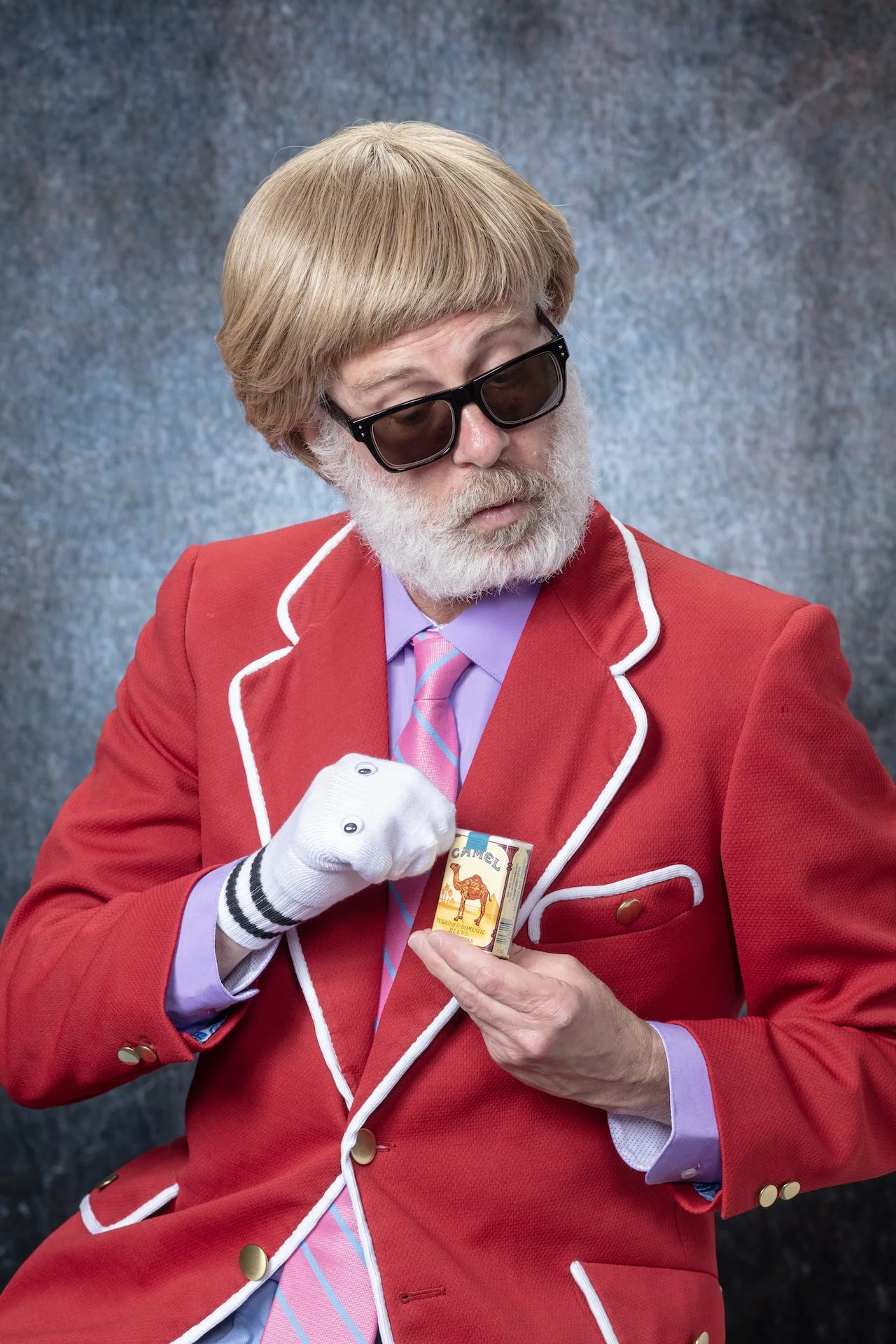 A man with a white beard and glasses wearing a red blazer with white trim, holding a pack of Camel cigarettes.