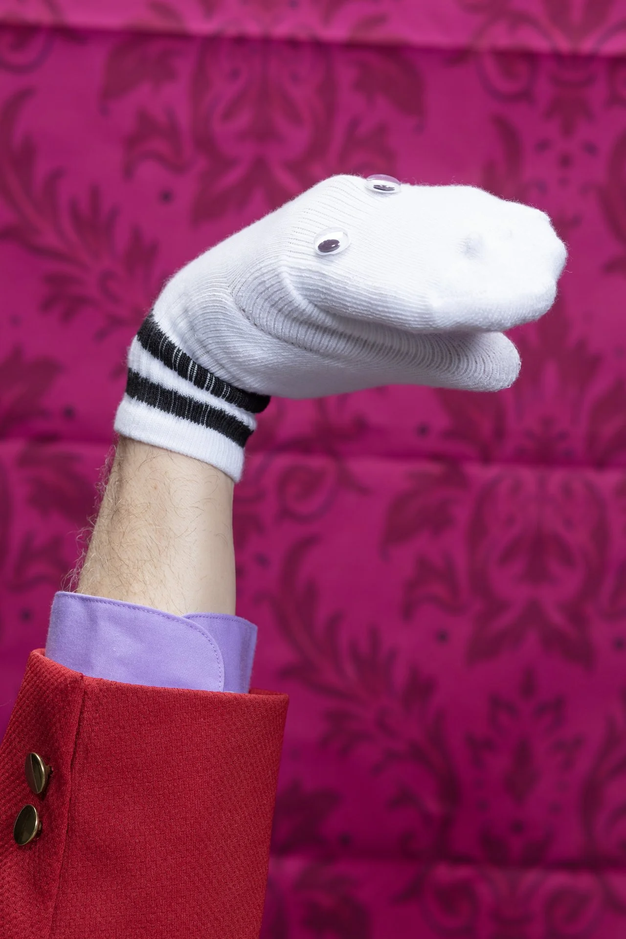 A hand wearing a white sock with googly eyes, pointing to the right, against a pink patterned background. The person is dressed in colorful clothing including a purple sleeve and a red coat.
