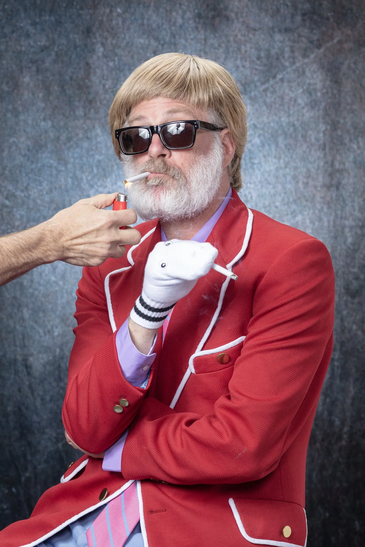 Man with gray beard wearing black sunglasses and a red blazer, lighting a cigarette with a lighter.
