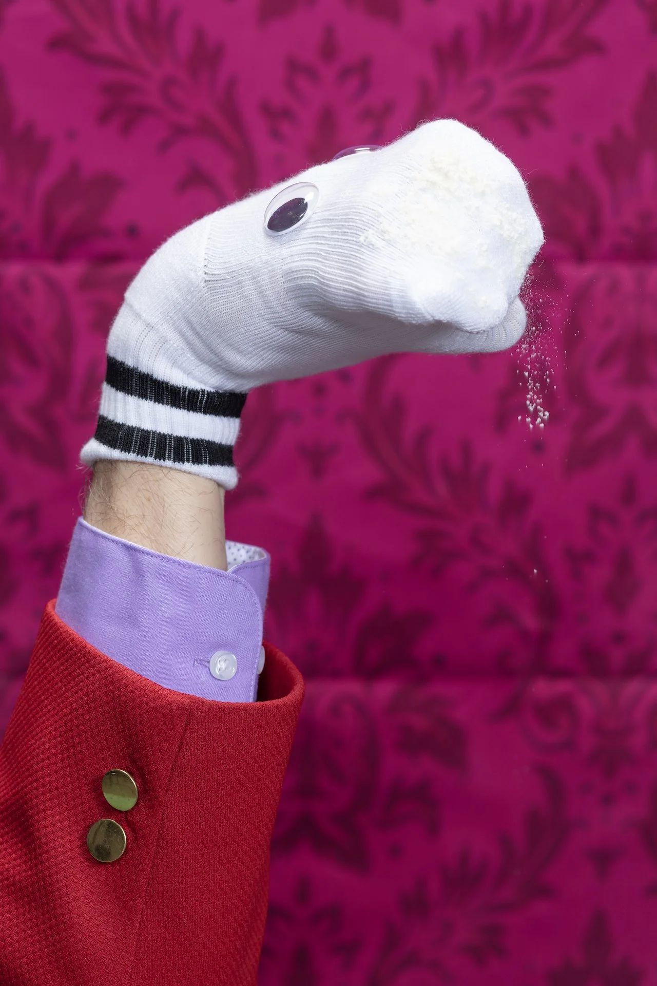 A puppet resembling a rat with googly eyes, wearing a white sock, being held in a person's hand, dressed in a red coat with gold buttons, against a bright pink textured background.