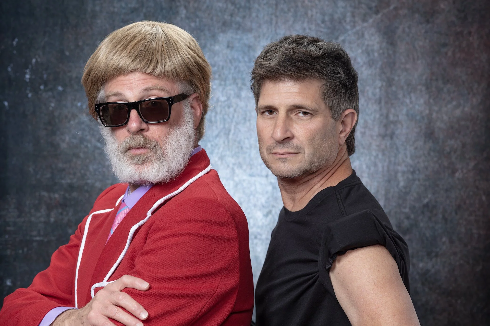 Two men pose back-to-back against a dark, textured background. The man on the left has a beard, wearing sunglasses, a red jacket, and a colorful shirt. The man on the right has short, dark hair, and is wearing a black T-shirt.