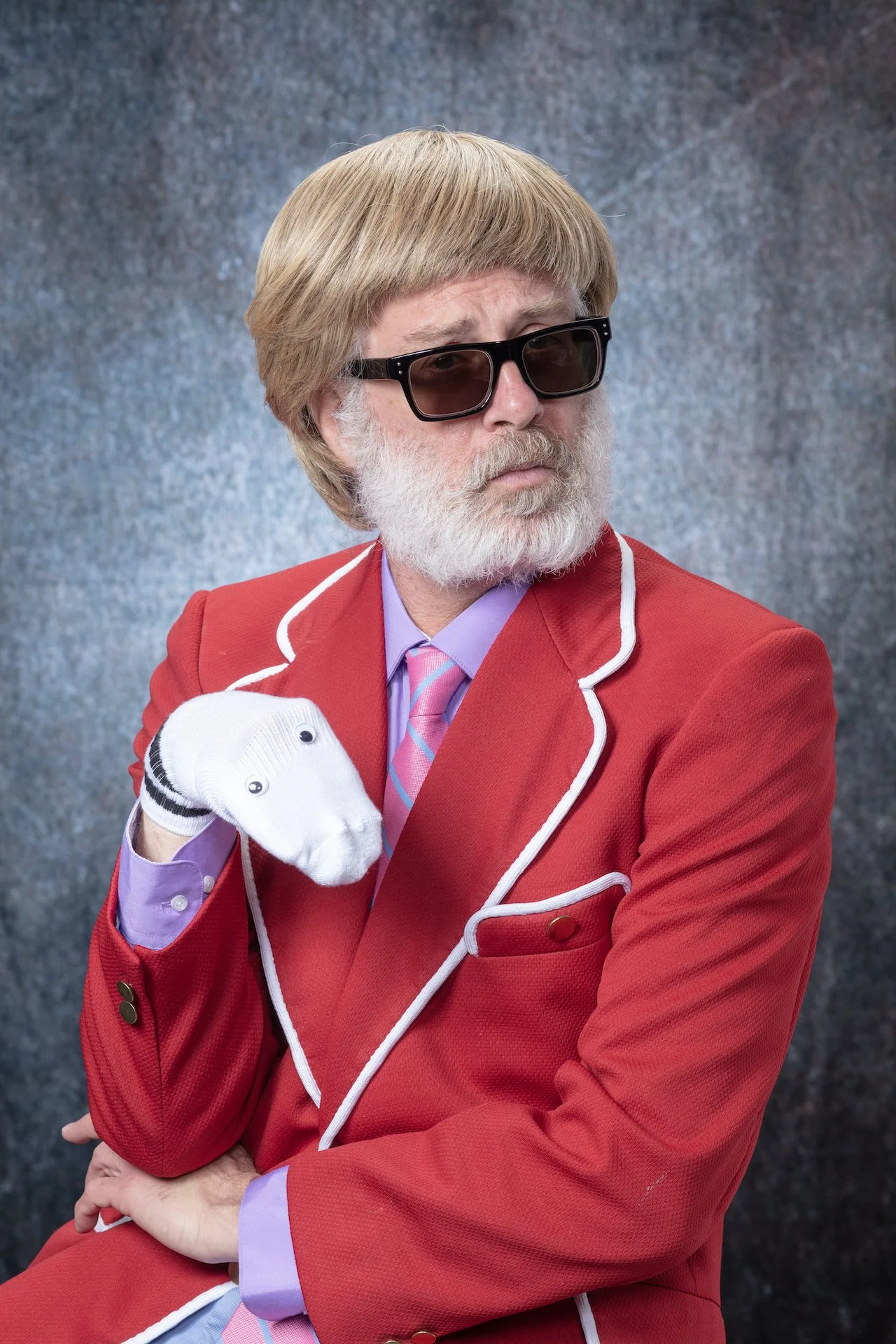 A person dressed as a golf enthusiast, wearing a red blazer with white piping, lavender shirt, pink striped tie, black sunglasses, and white golf glove, holding a golf club over their shoulder against a gray textured background.
