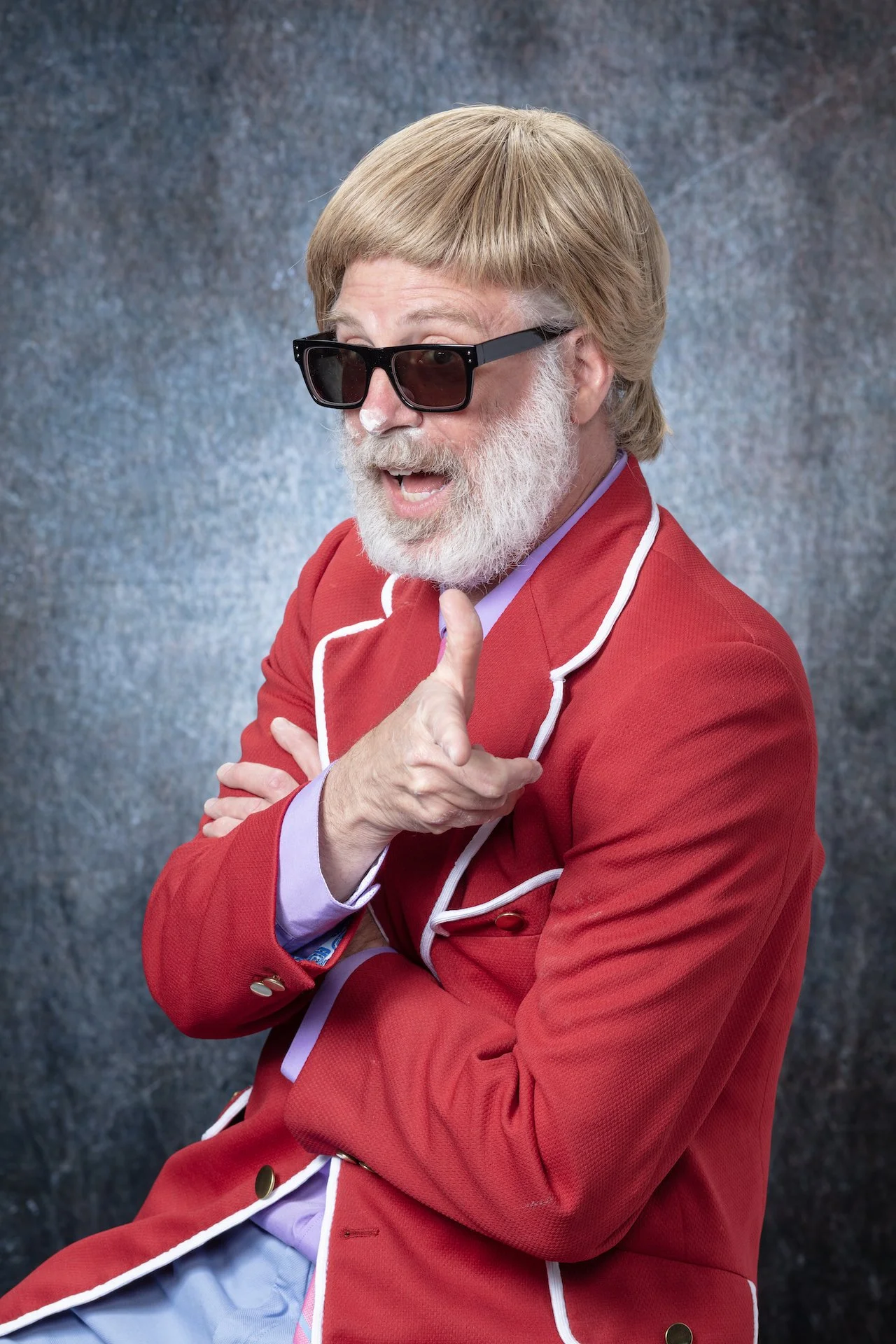 A man with a gray beard, wearing black sunglasses, a red coat with white piping, and a purple shirt, pointing with his right hand and smiling, against a textured blue and gray background.