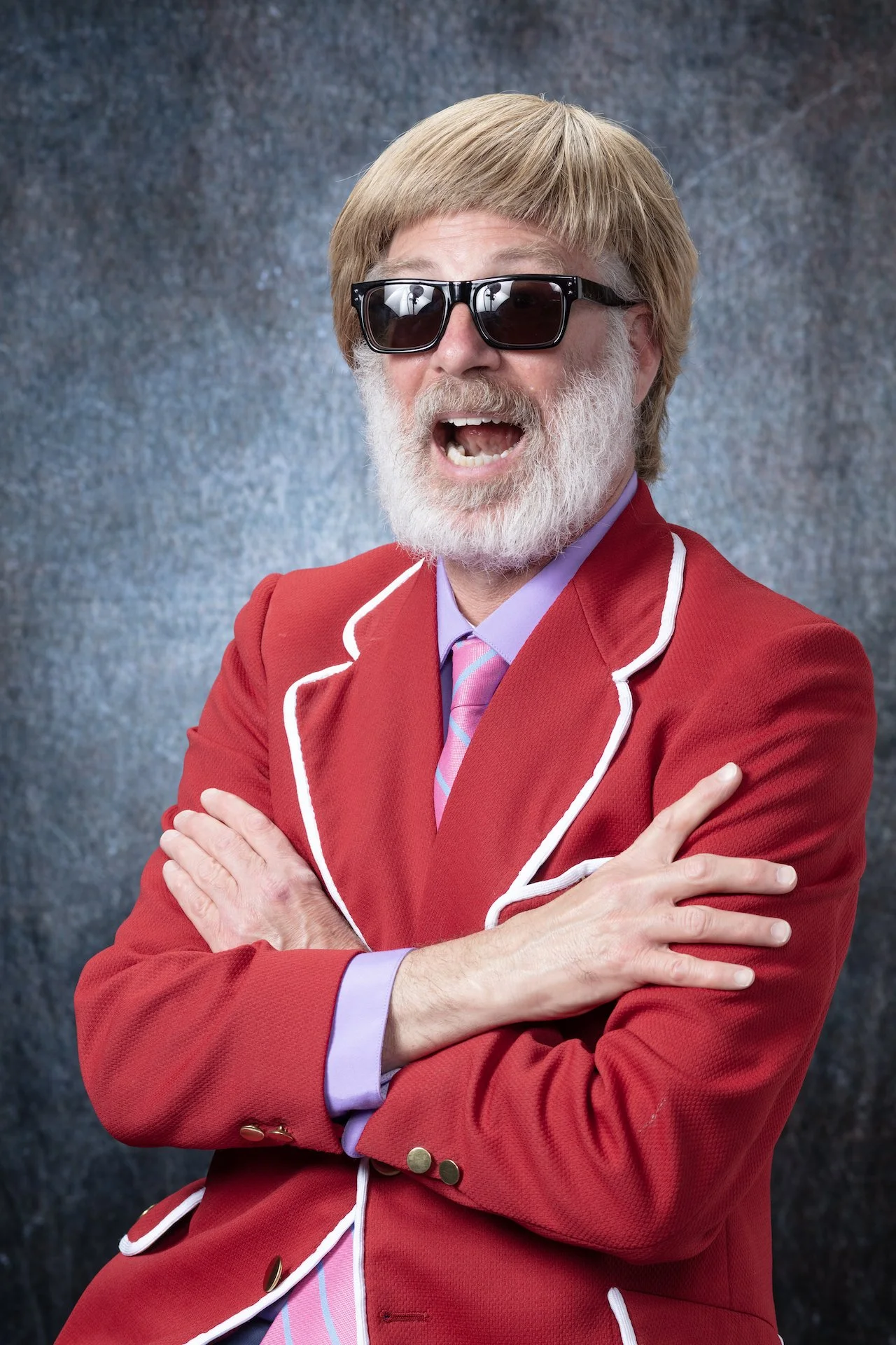A man with a beard and mustache wearing black sunglasses, a red blazer with white piping, a lavender shirt, and a pink striped tie. He is smiling with his mouth open and arms crossed over his chest, against a textured gray backdrop.