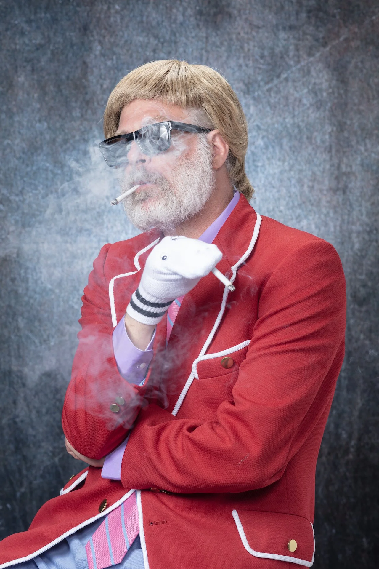A man with a beard wearing a red blazer with white piping, a purple dress shirt, and a striped tie, smoking a cigarette and wearing sunglasses. Smoke surrounds him against a textured background.