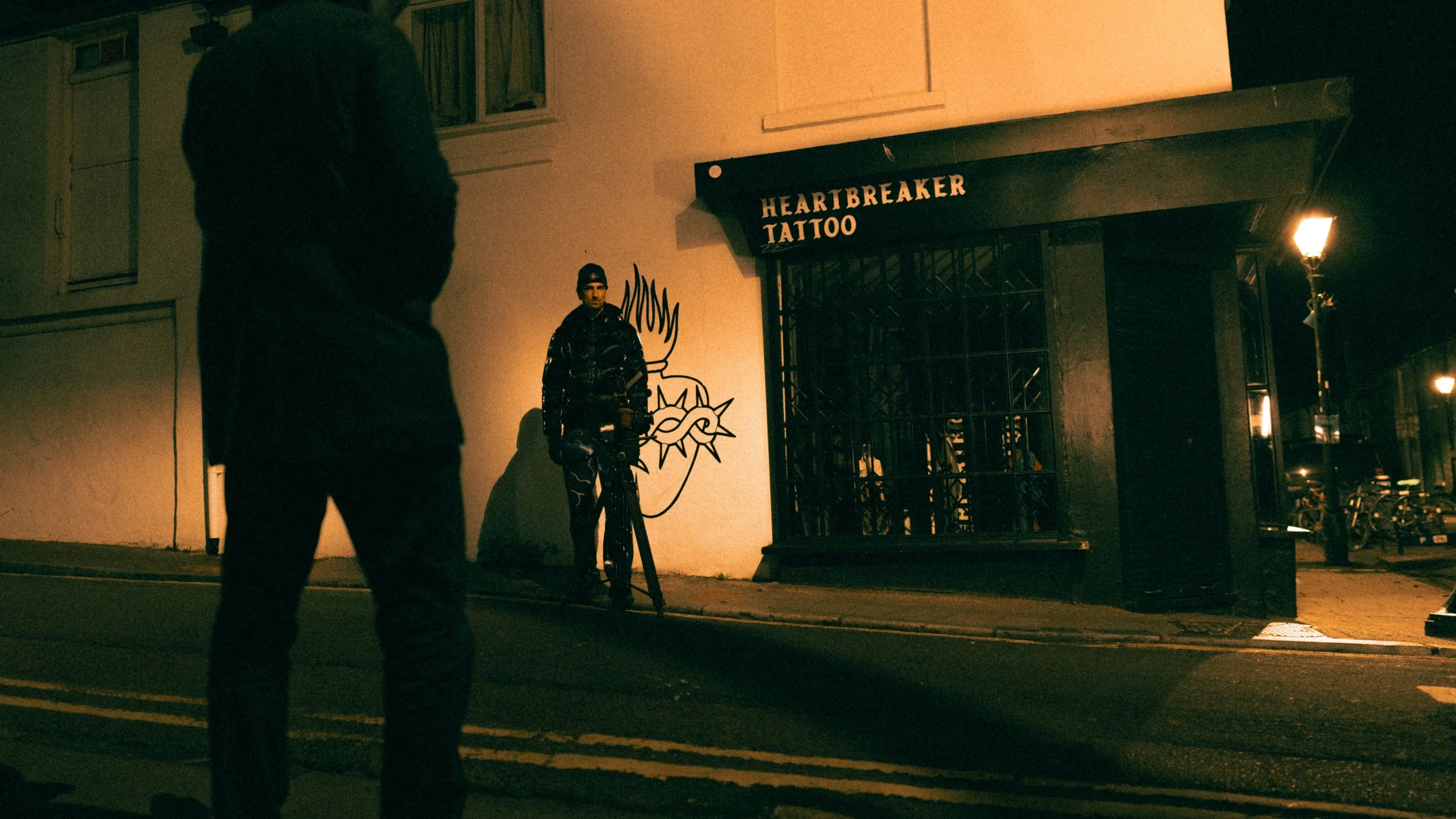A person standing on a dimly lit street at night facing another person standing near a graffiti art depiction of a human face. The building behind has a sign that says 'Heartbreaker Tattoo'.