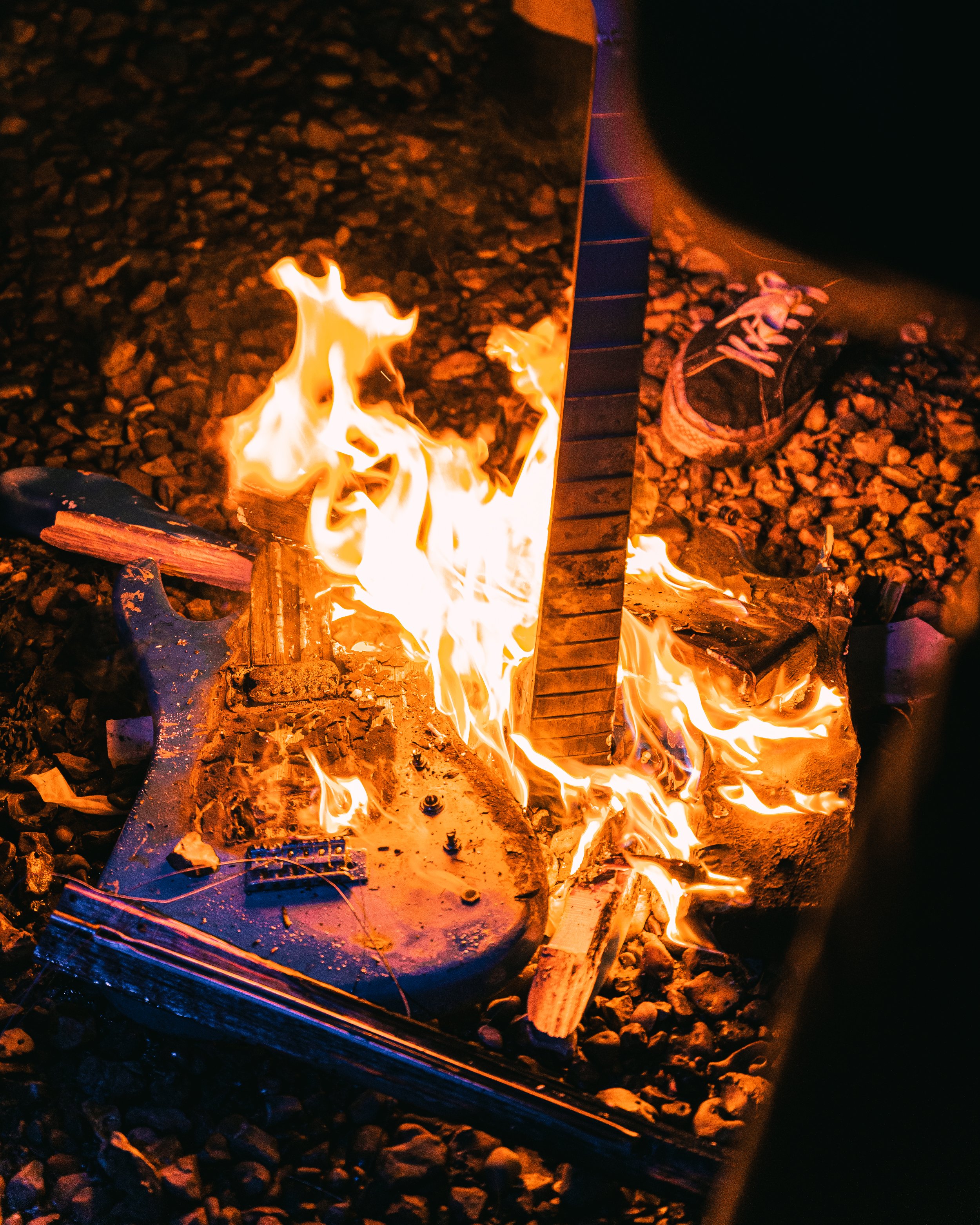 An electric guitar catching fire on a gravel surface, with one sneaker visible nearby.