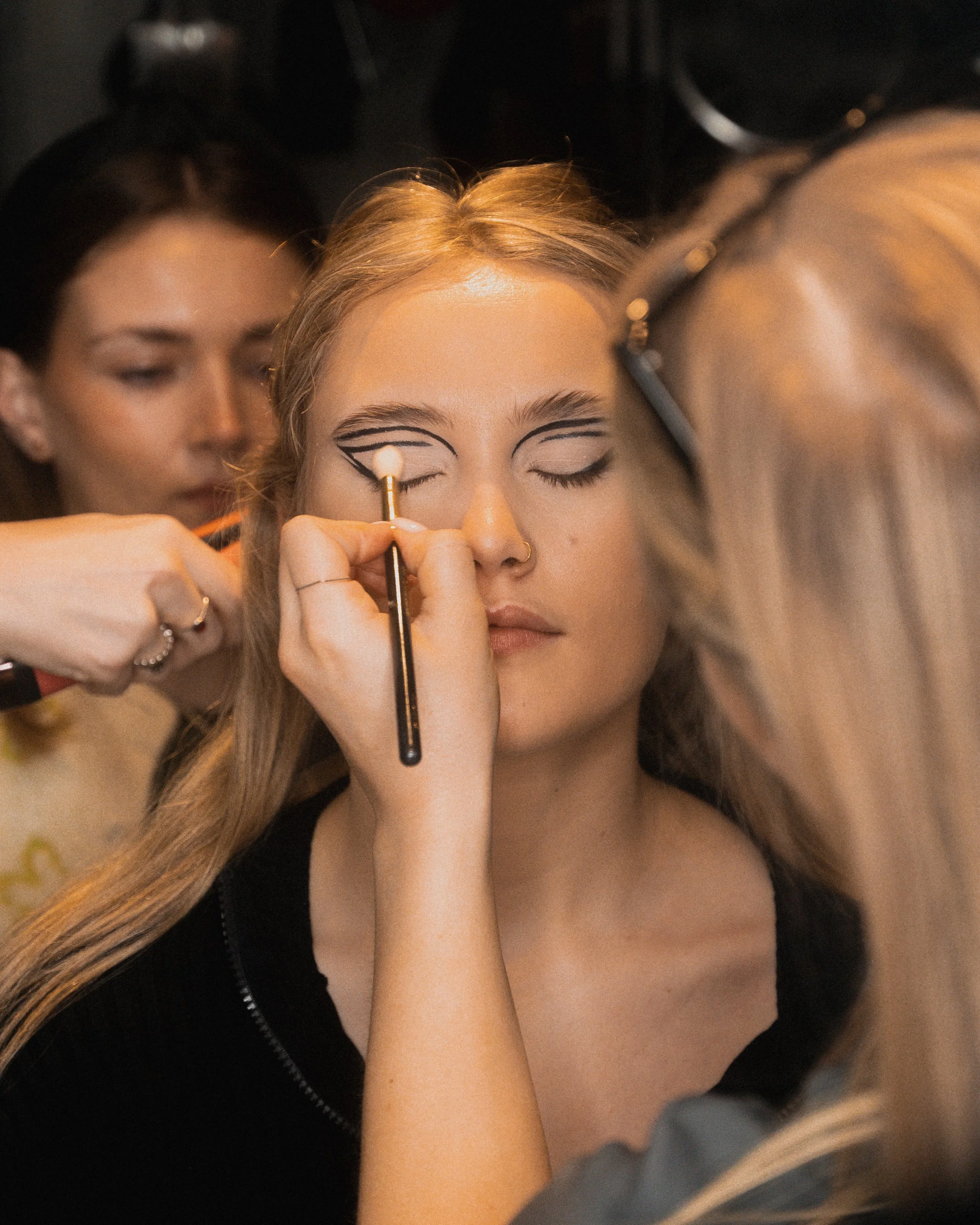 A woman with her eyes closed is getting makeup applied by a makeup artist, who is drawing a graphic design around her eyes for a look or costume. Another woman is in the background.