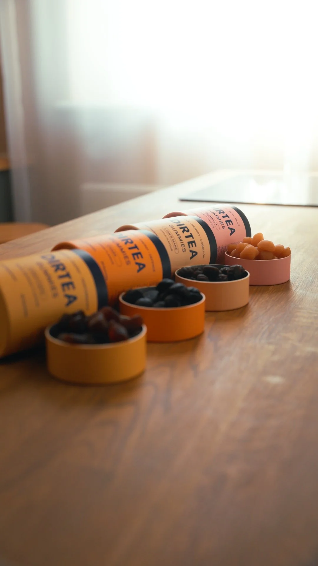 Three orange tubes labeled 'Gurtea Gummies' and three small round containers filled with gummies or candies in black, brown, and yellow colors on a wooden table near a window.