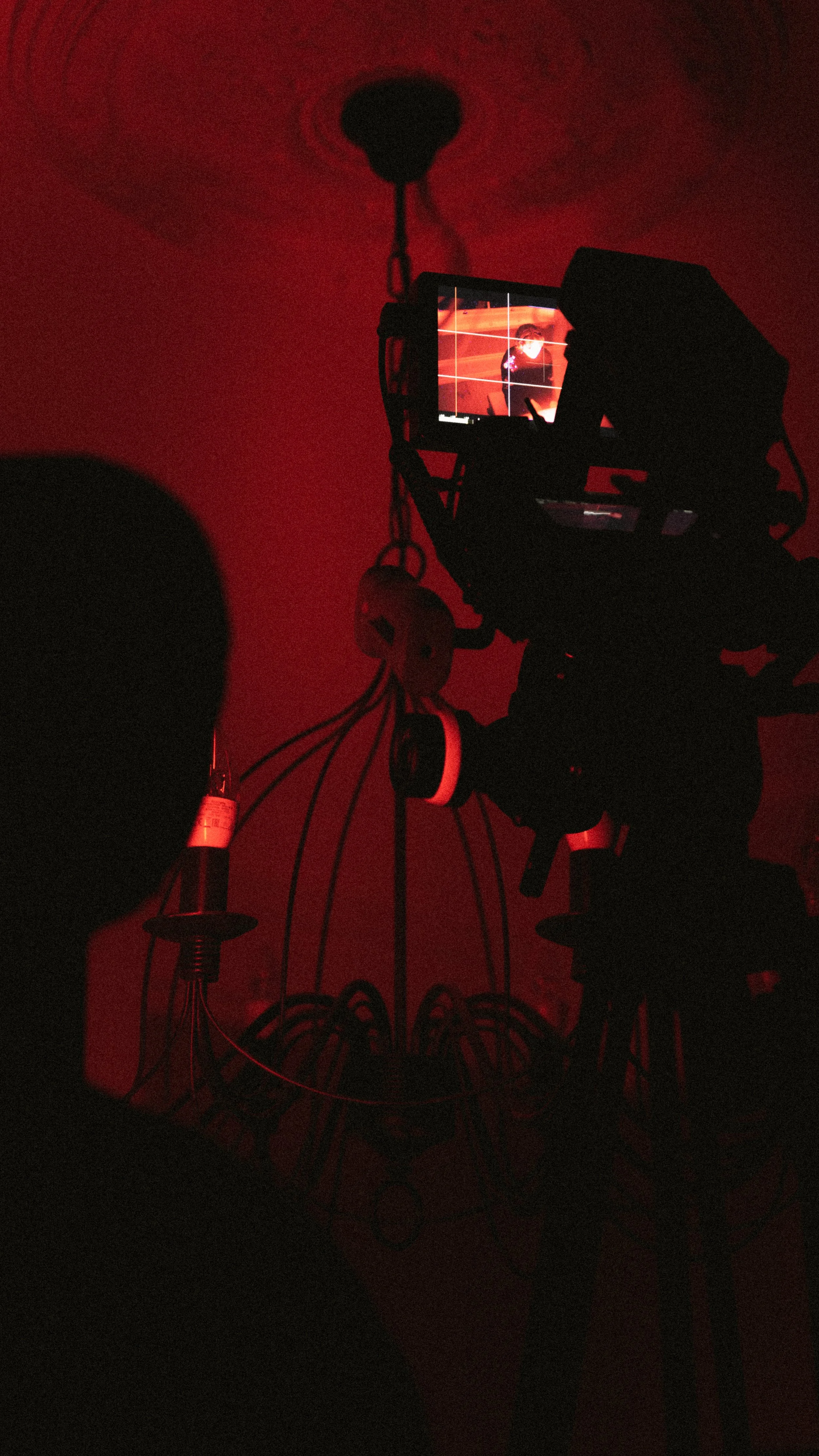 A camera on a tripod captures a scene with a red light illuminating a chandelier and a person wearing a hood, creating silhouettes and a moody atmosphere.