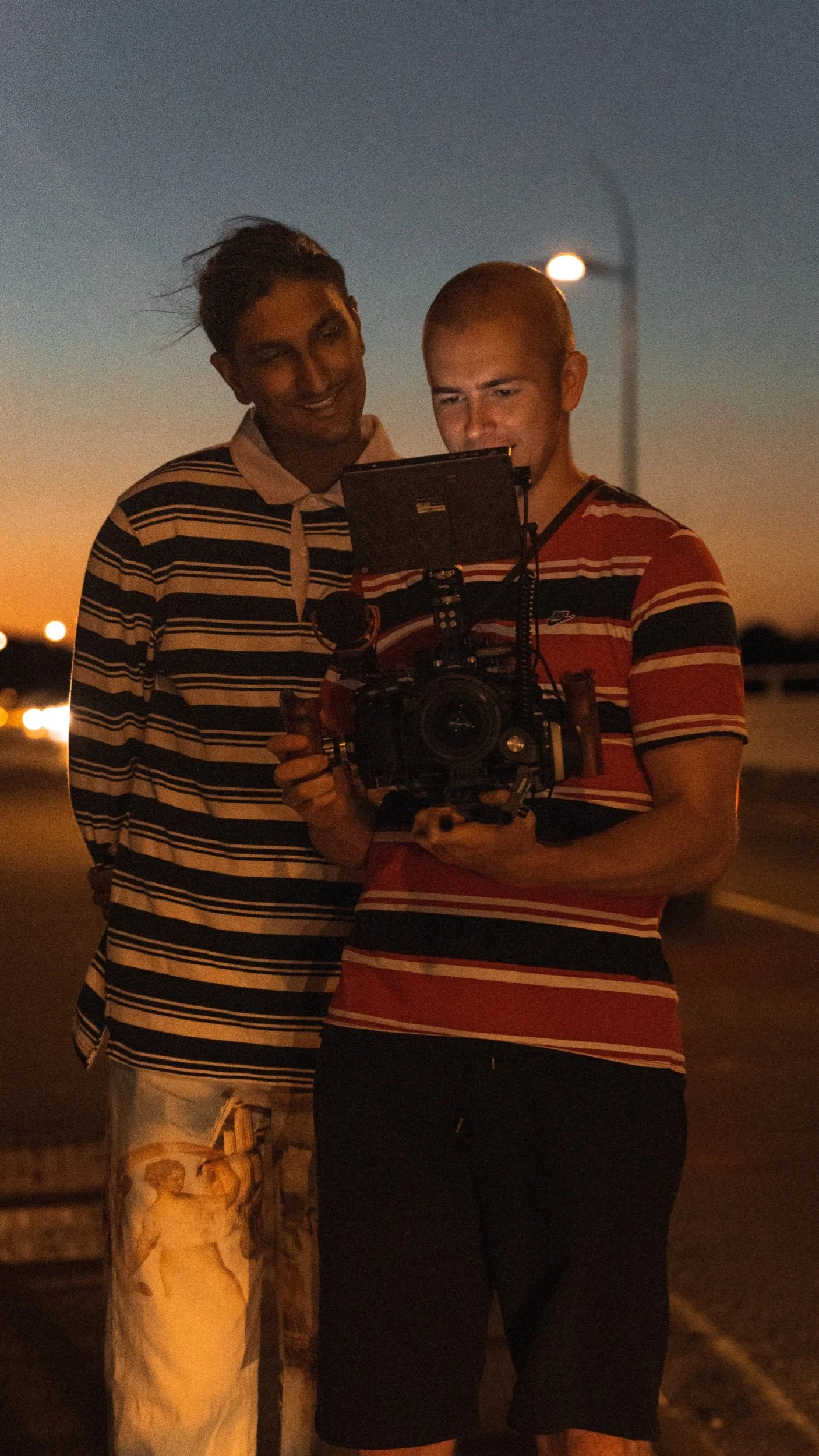 Two young men stand together outdoors at dusk, looking at a camera's screen.