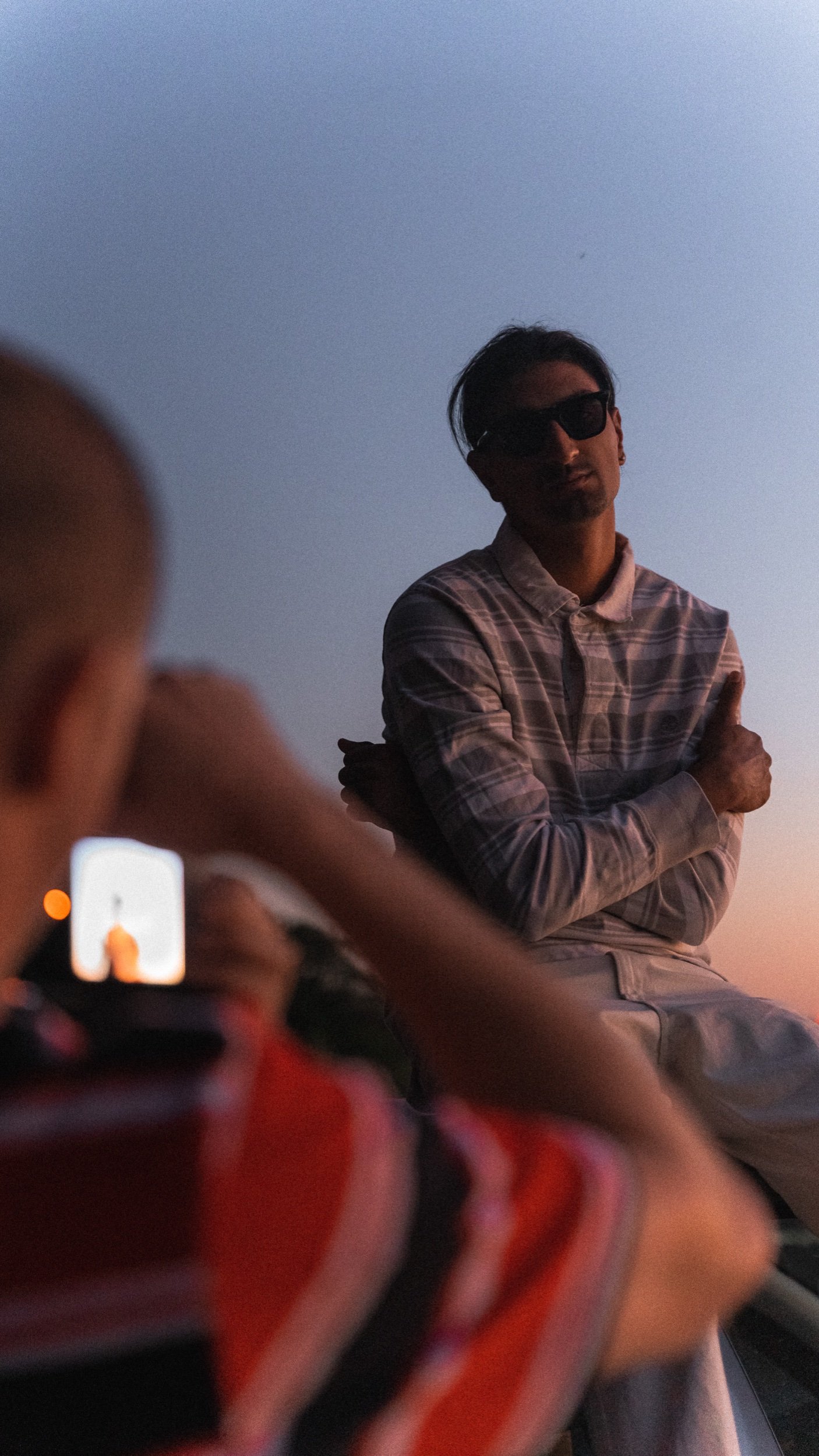 A man with dark hair and sunglasses sitting outdoors during sunset, with two people in the foreground taking pictures of him.