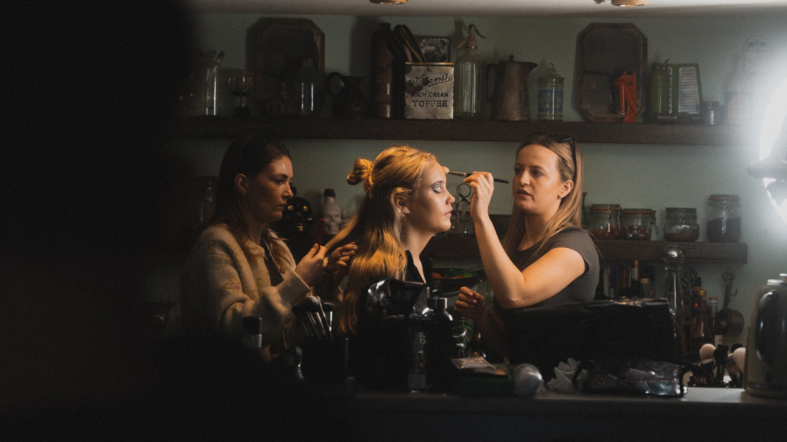 Three women are in a kitchen, with one applying makeup to another, while the third looks on. The scene is illuminated from the right side, with shelves and various kitchen items in the background.