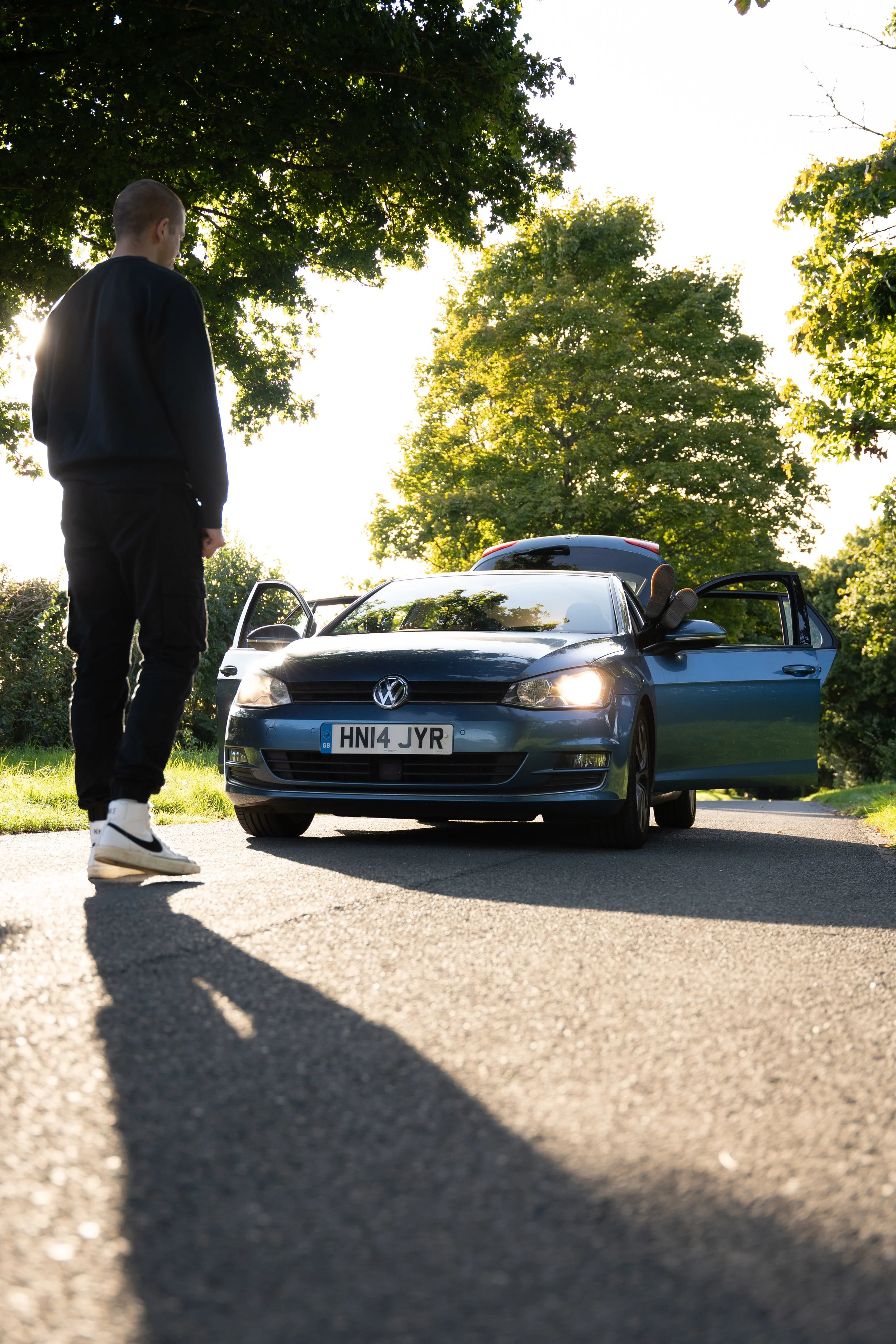 A young man standing on a quiet road with a blue Volkswagen car parked nearby. The car's door is open, and the sun is shining through trees, creating shadows on the pavement.