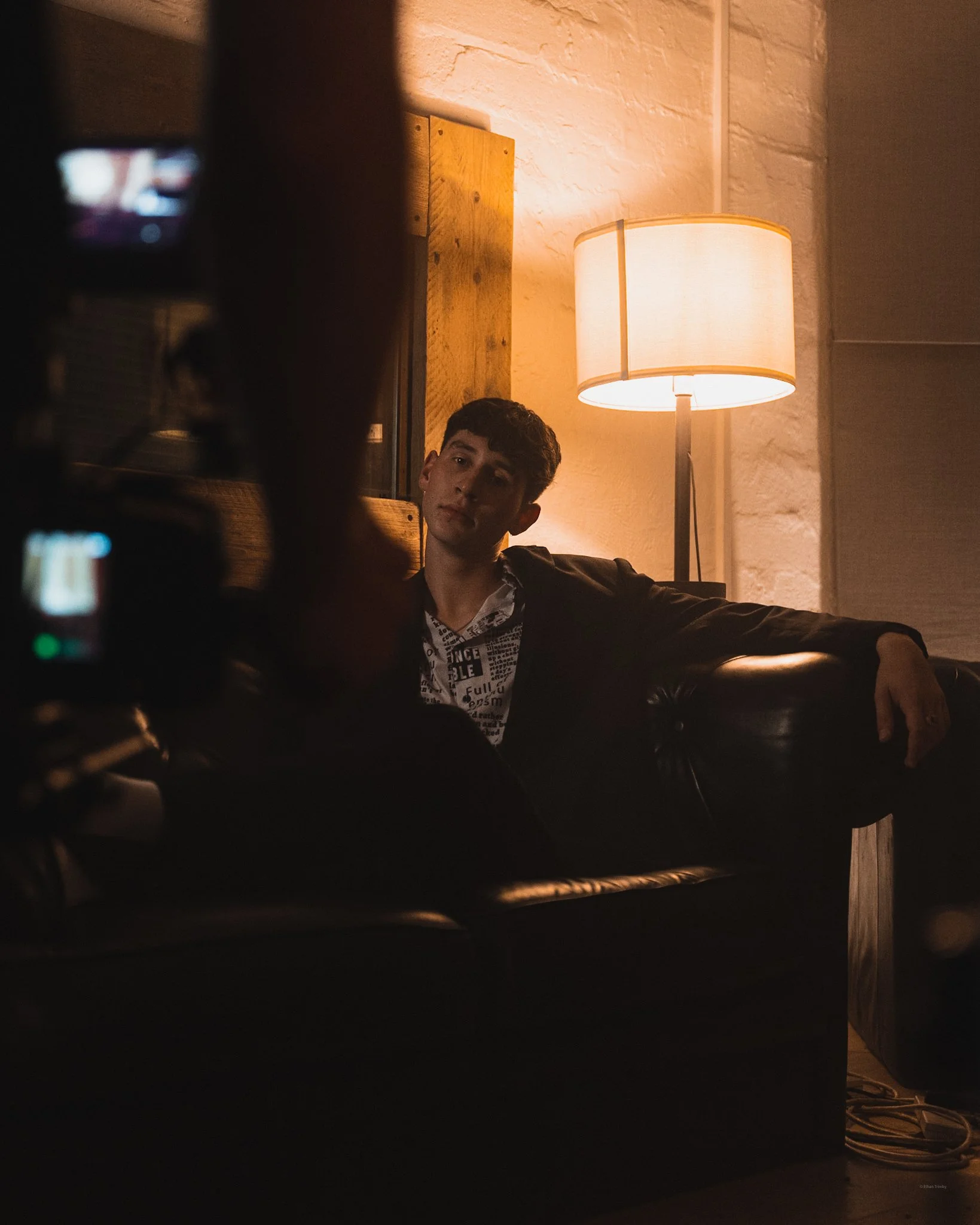 A young man sitting on a black leather couch in a dimly lit room with a floor lamp behind him. The room has white brick walls and wooden accents, creating a cozy atmosphere. Someone is taking a photo of him.