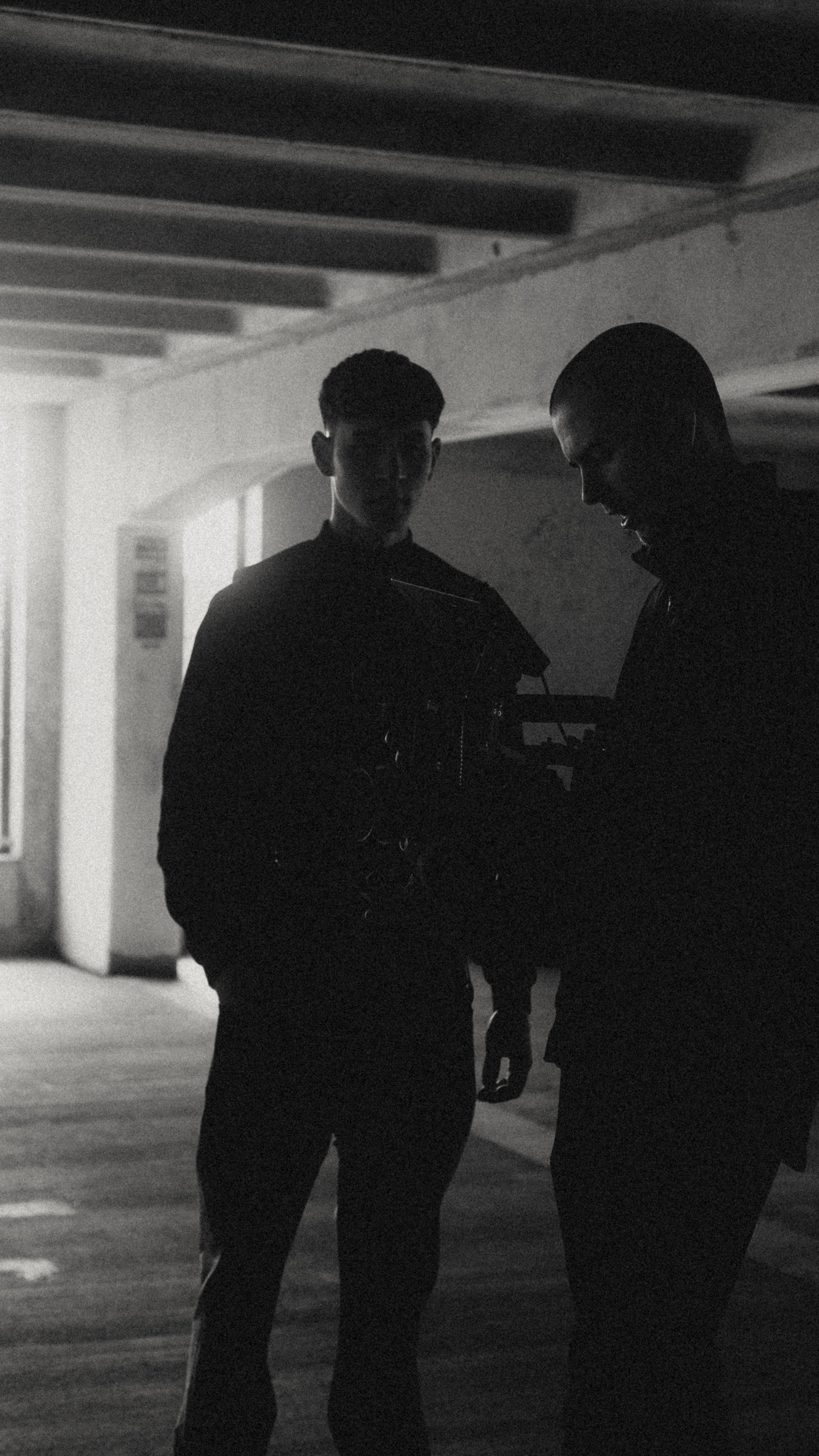 Two young men in dark clothing standing in an urban underpass, engaged in conversation in low lighting.