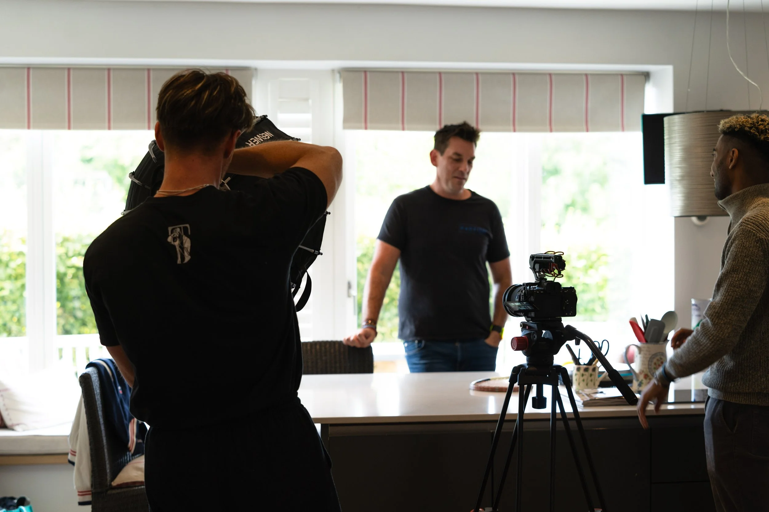 Three men preparing for a video shoot in a bright room with large windows and natural light. One man is adjusting a camera on a tripod, another is standing near a table filled with various items, and the third man is standing in front of the camera, 
