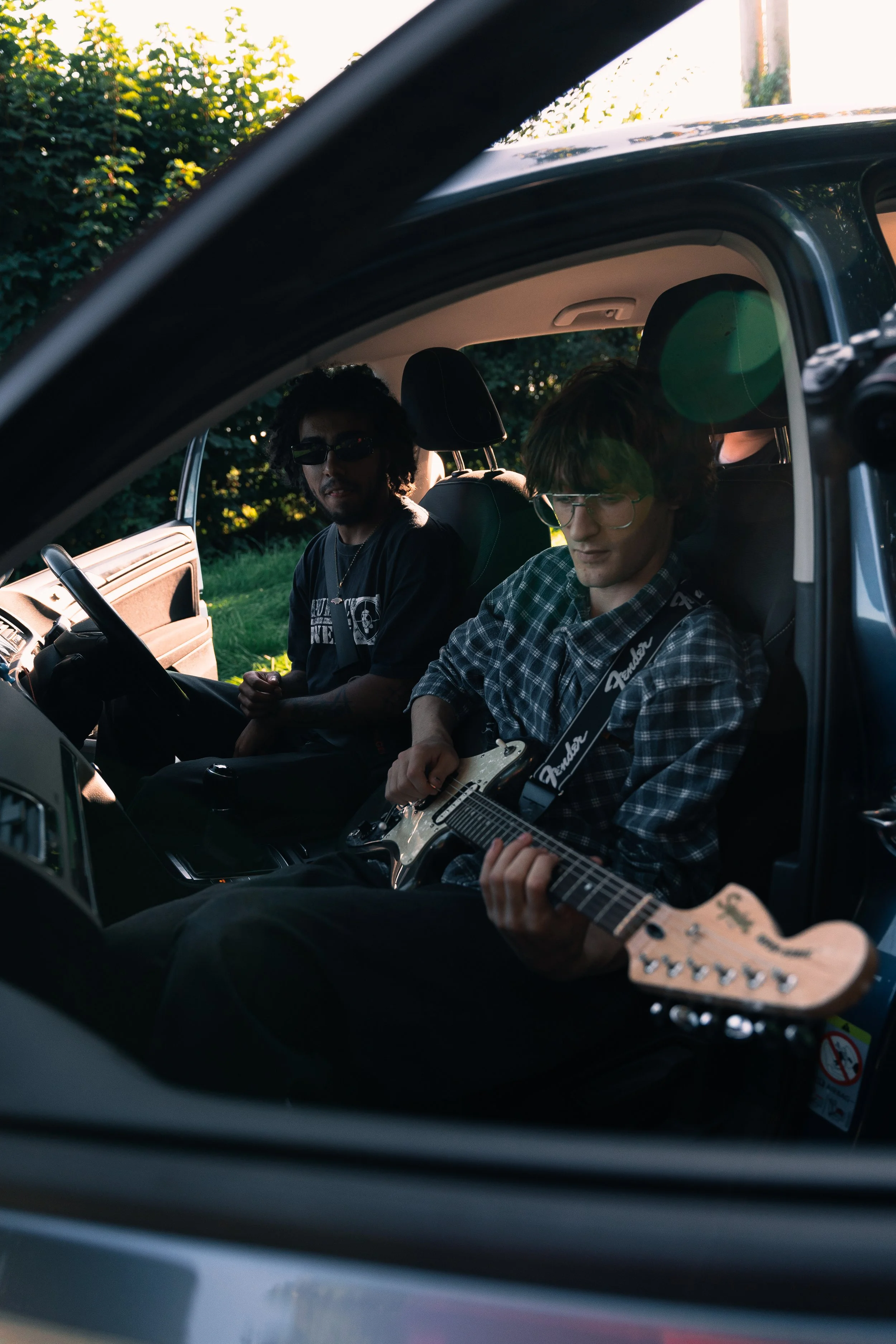 Two young men sitting inside a car, one playing an electric guitar, during a sunny day.