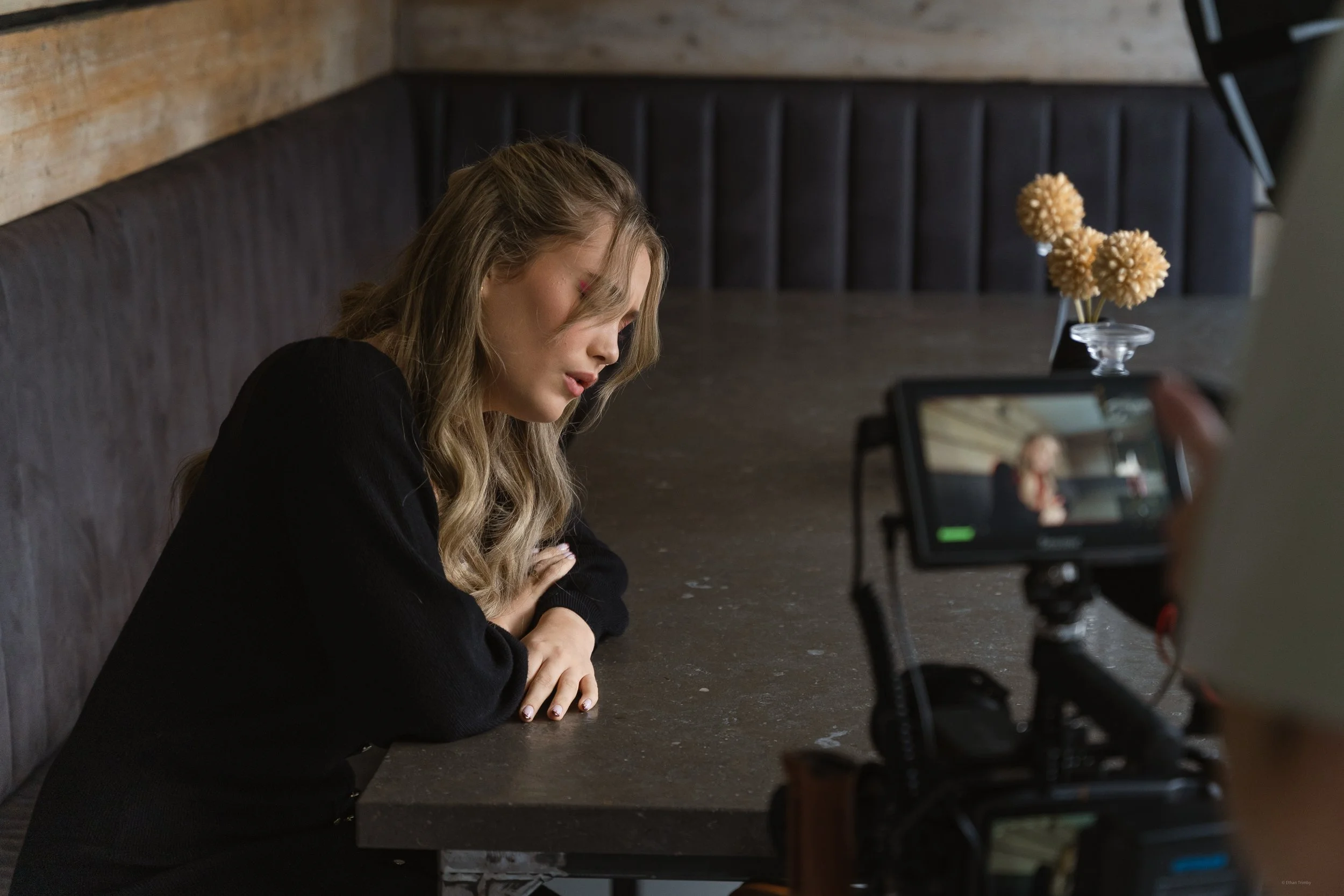 A woman with wavy blonde hair and a black shirt is sitting at a table with her head resting on her crossed arms, appearing to be in emotional distress during a video recording or interview.