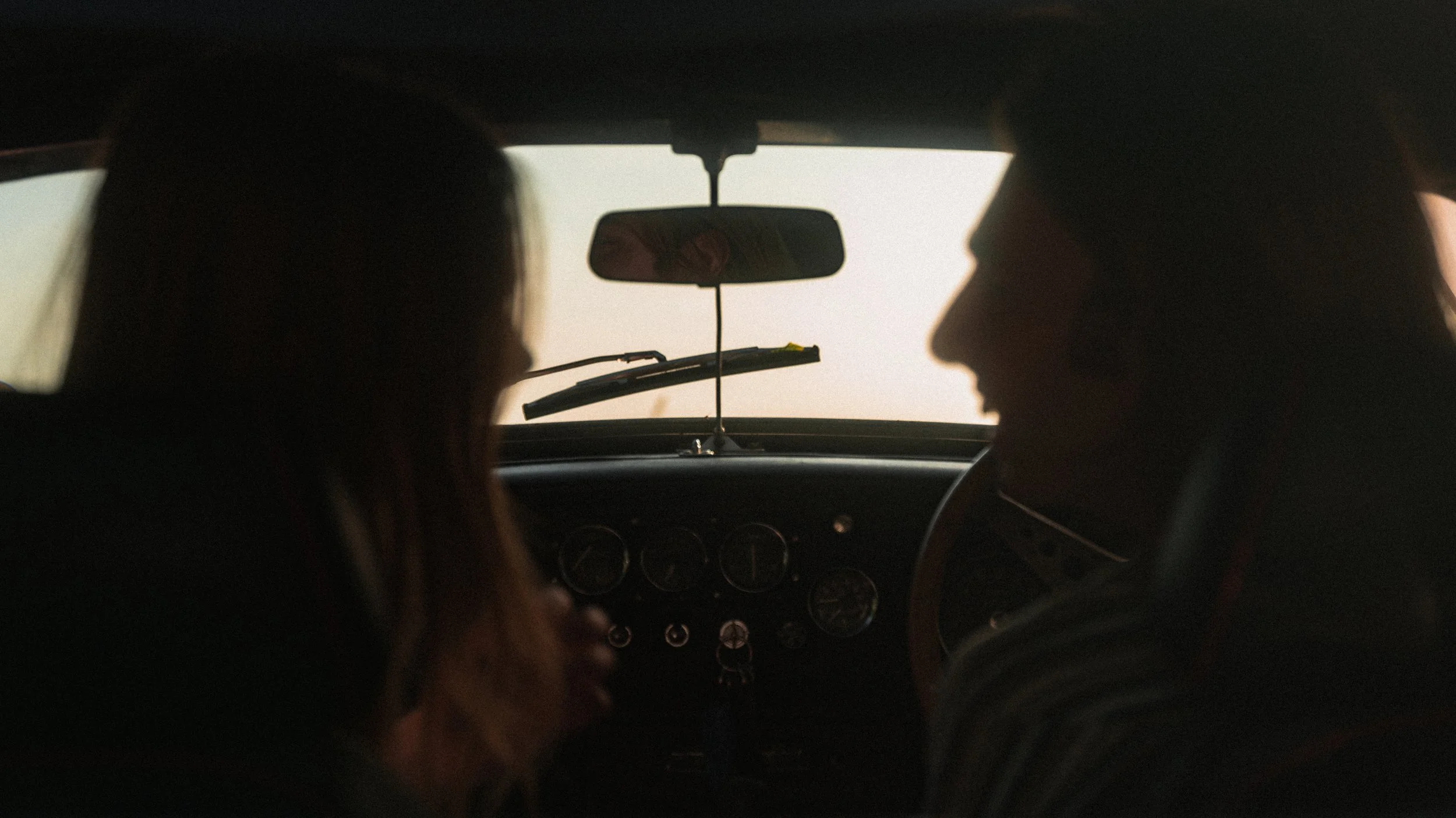 Silhouettes of two people sitting inside a vintage car, facing each other, with a blurred view of the dashboard and windshield.