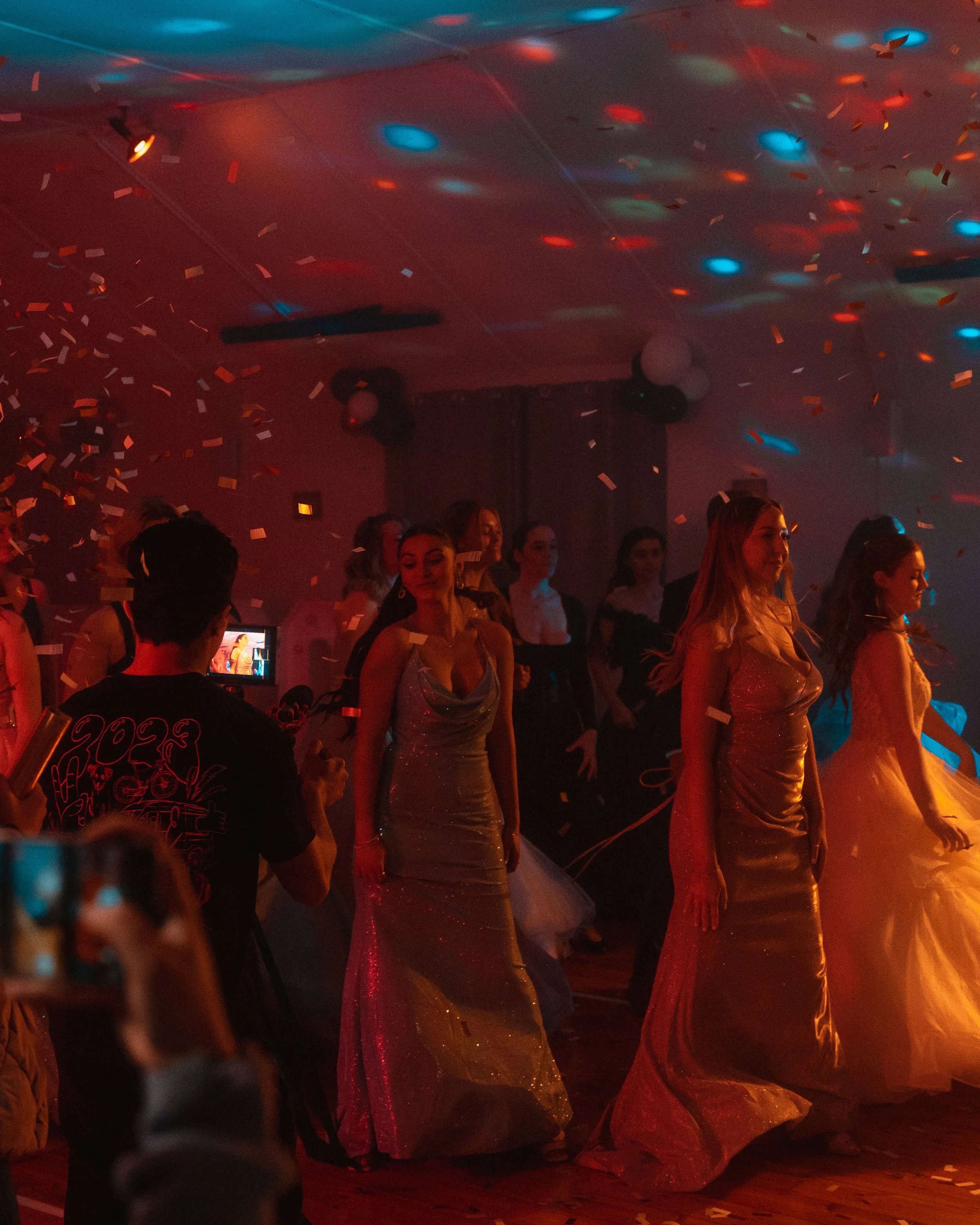 Women in formal gowns dancing at a party with colorful lighting and confetti in the air.