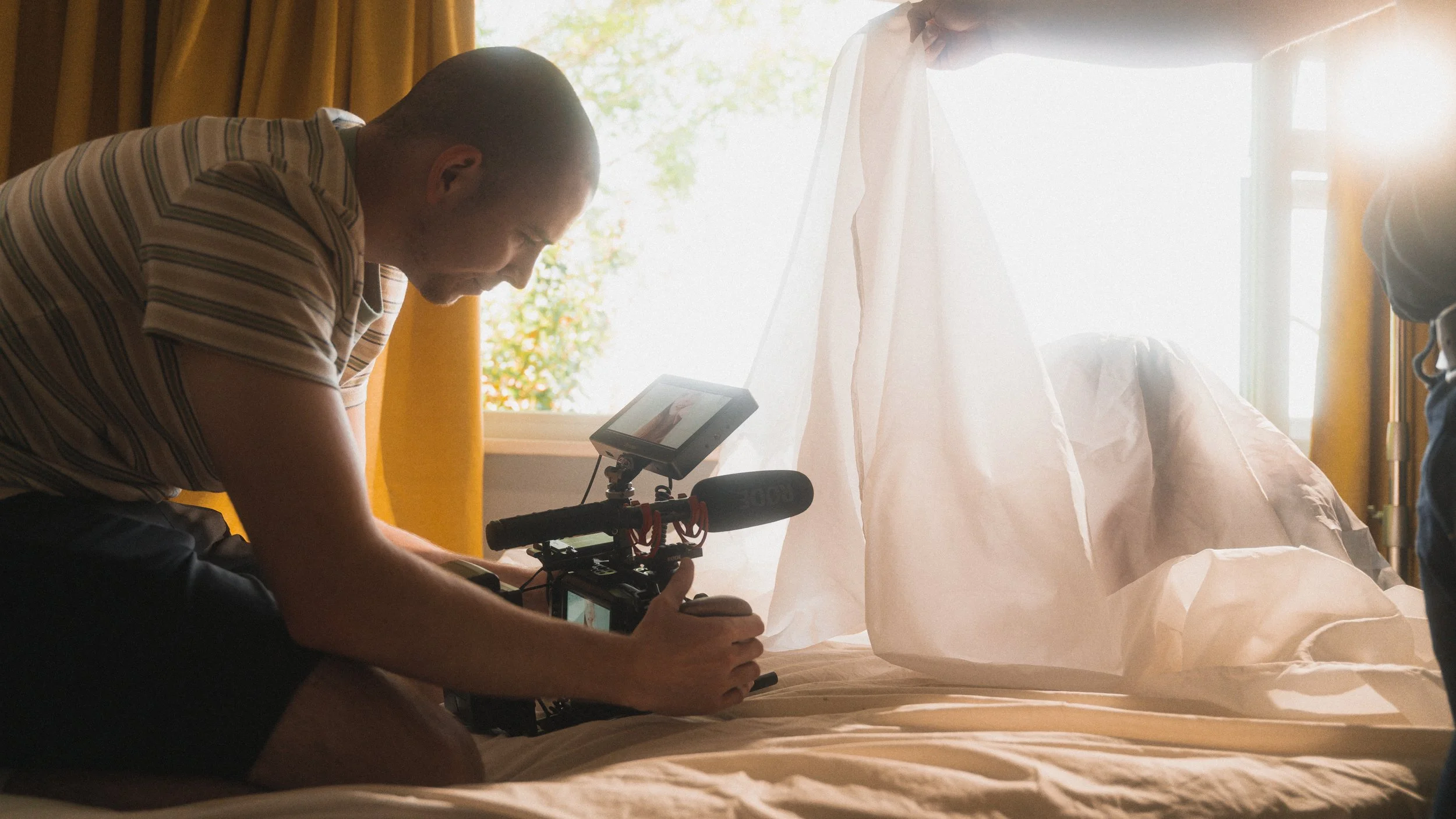 A person operating a camera on a bed, filming a bed with white sheets and a partially covered person with a sheet, in a sunlit room with yellow curtains