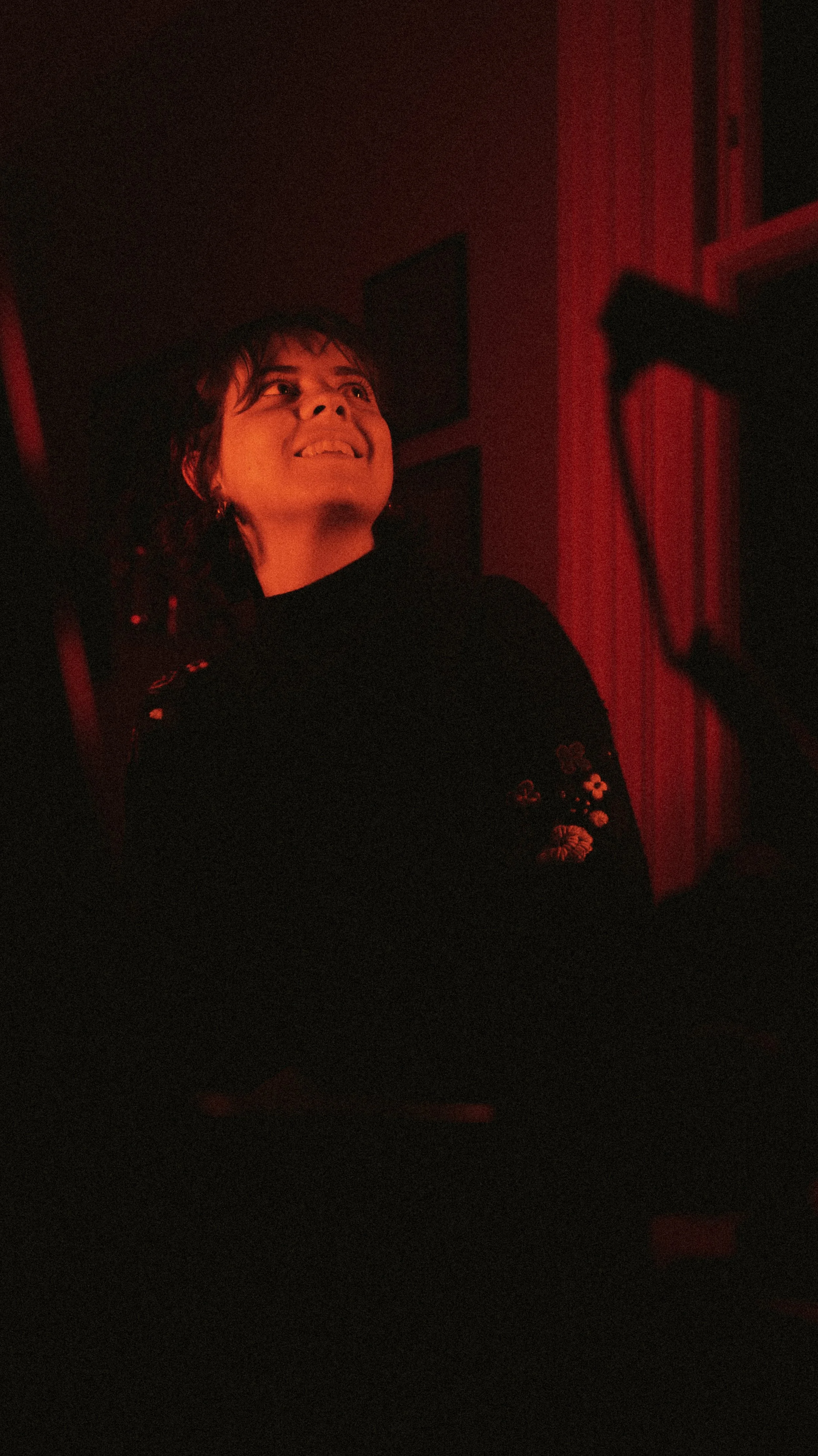 A woman with short hair smiling, illuminated by red lighting, sitting indoors near a keyboard or musical instrument.