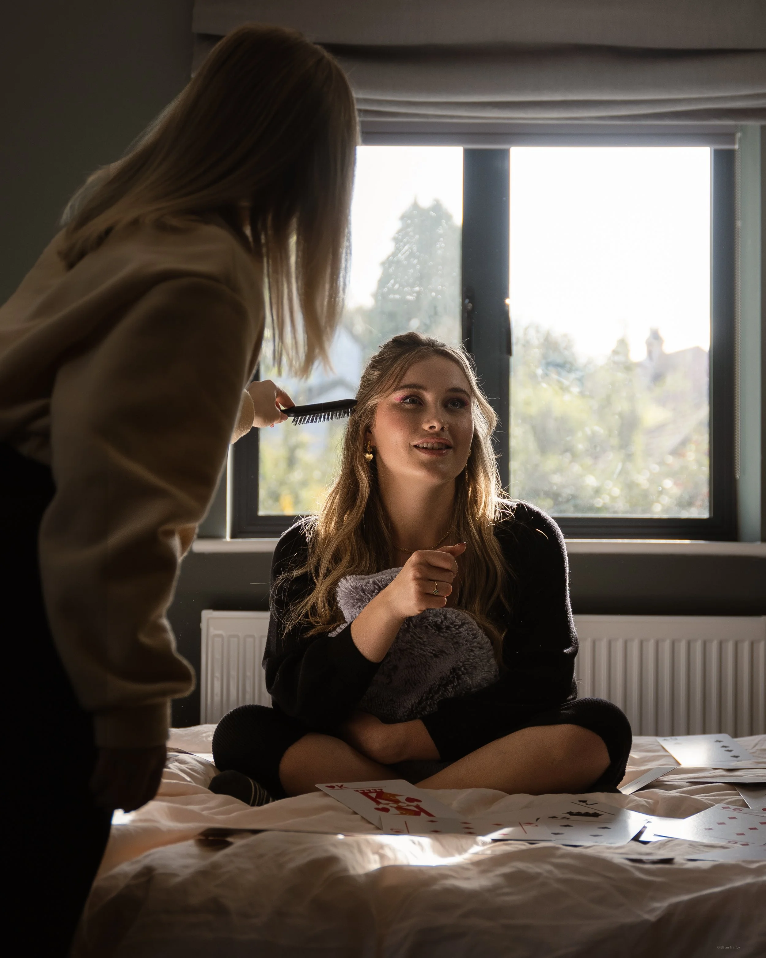 A young woman sitting on a bed with playing cards, having her hair done by another person in a room with a large window and natural daylight.