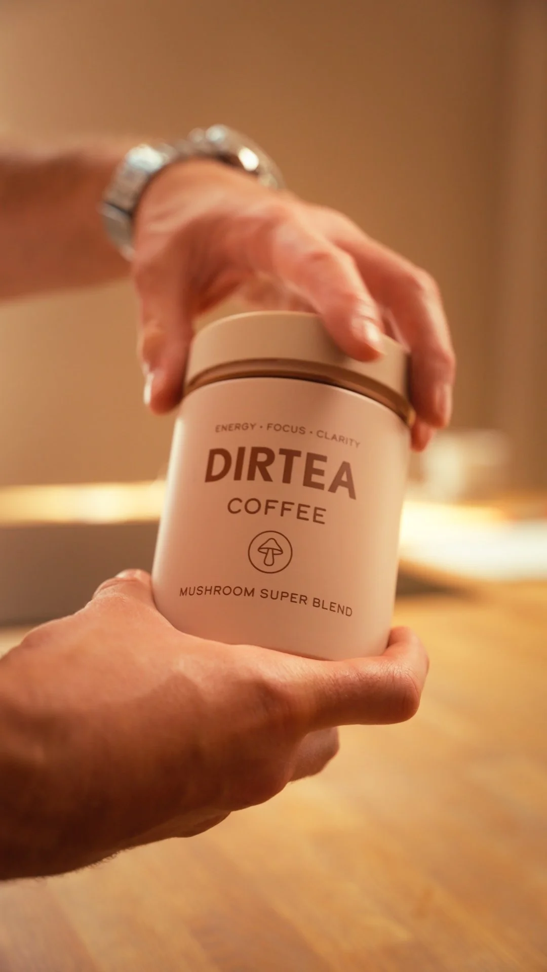 Close-up of a hand holding a container of DIRTÉA coffee with a mushroom logo, with another hand reaching to open it. The container is labeled 'energy, focus, clarity' and 'mushroom super blend'.