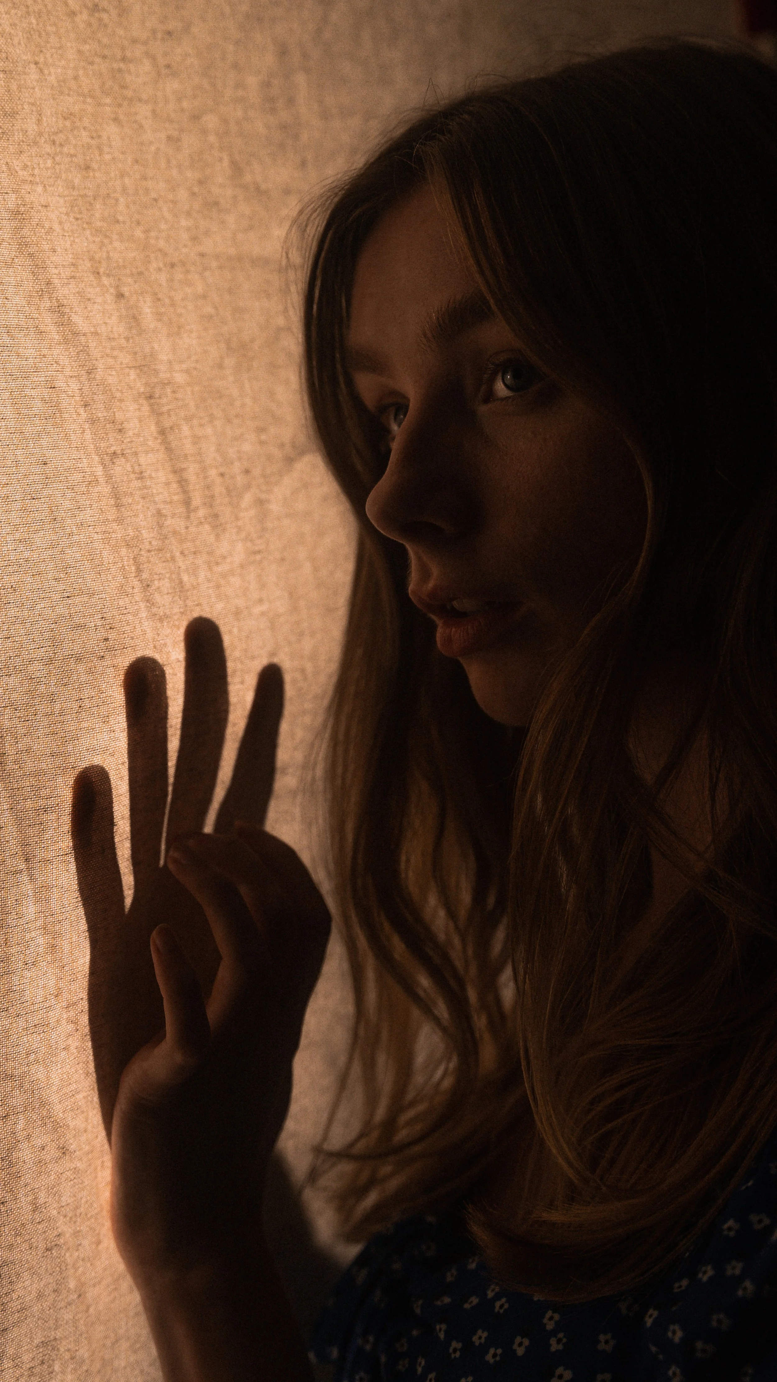 A young woman with long hair touching a textured wall, illuminated softly from the side, creating shadows of her hand on the wall.