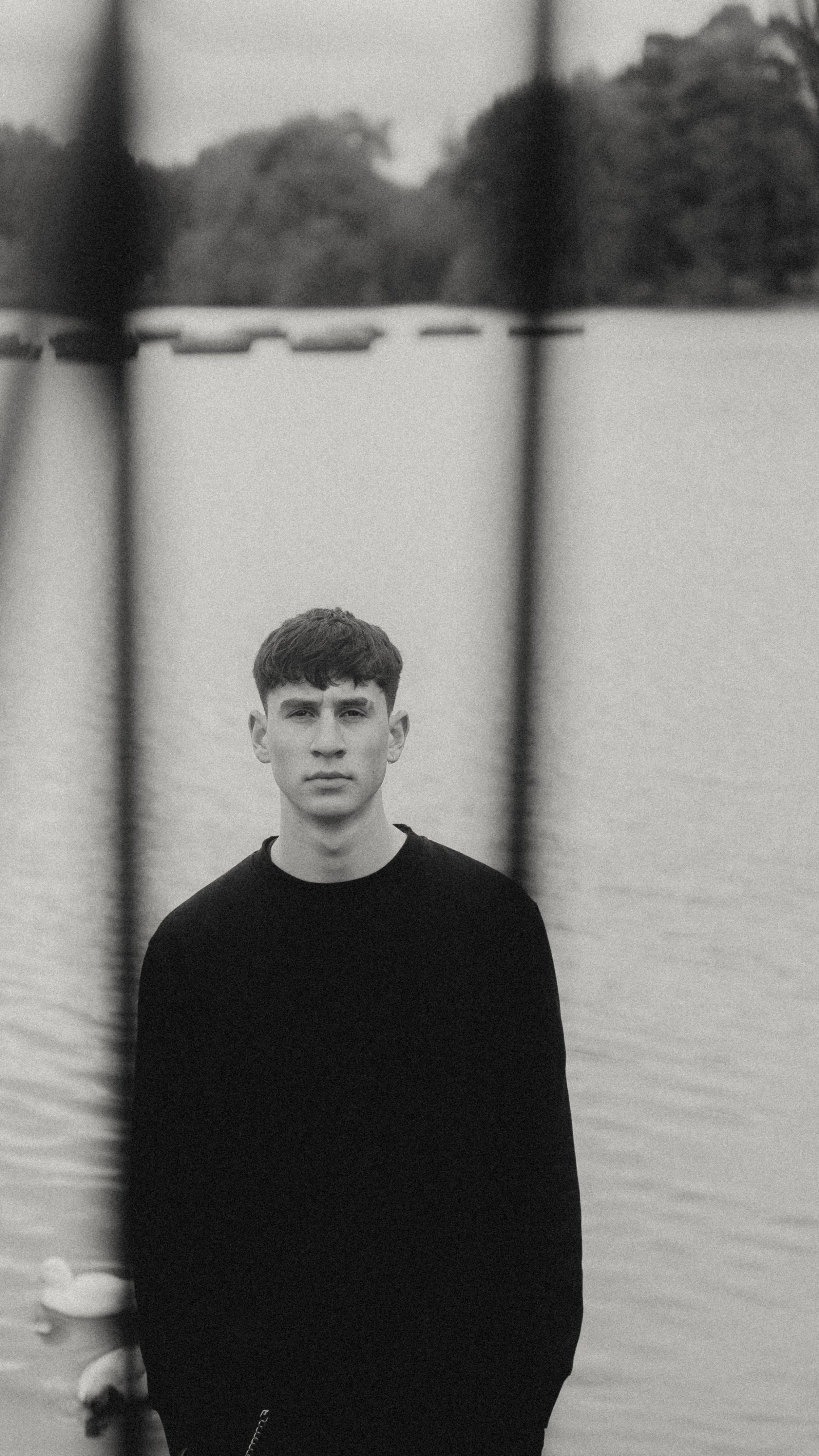 Black and white photo of a young man with short hair standing outdoors on a body of water, with trees in the background. The photo is taken through what appears to be a fence or barrier.
