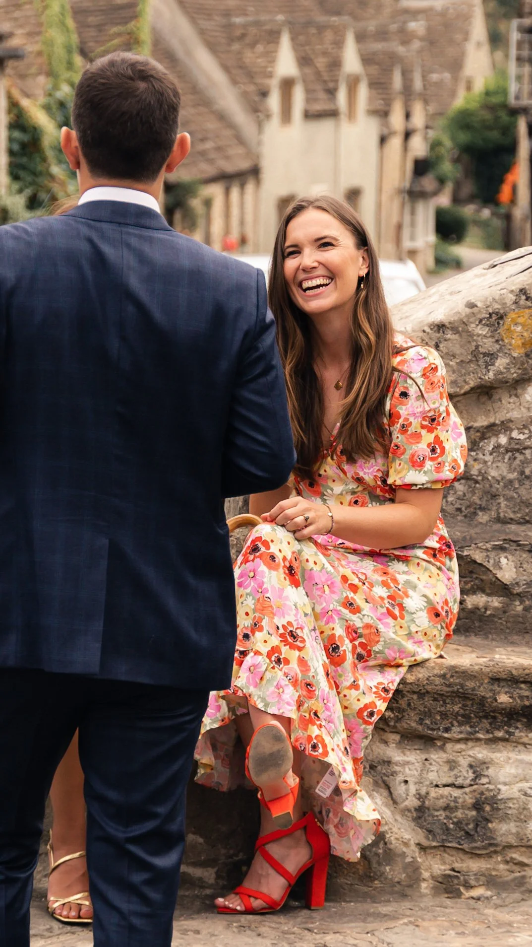 A woman in a colorful floral dress and red high heels, smiling as a man in a blue suit proposes or gives a ring outdoors.