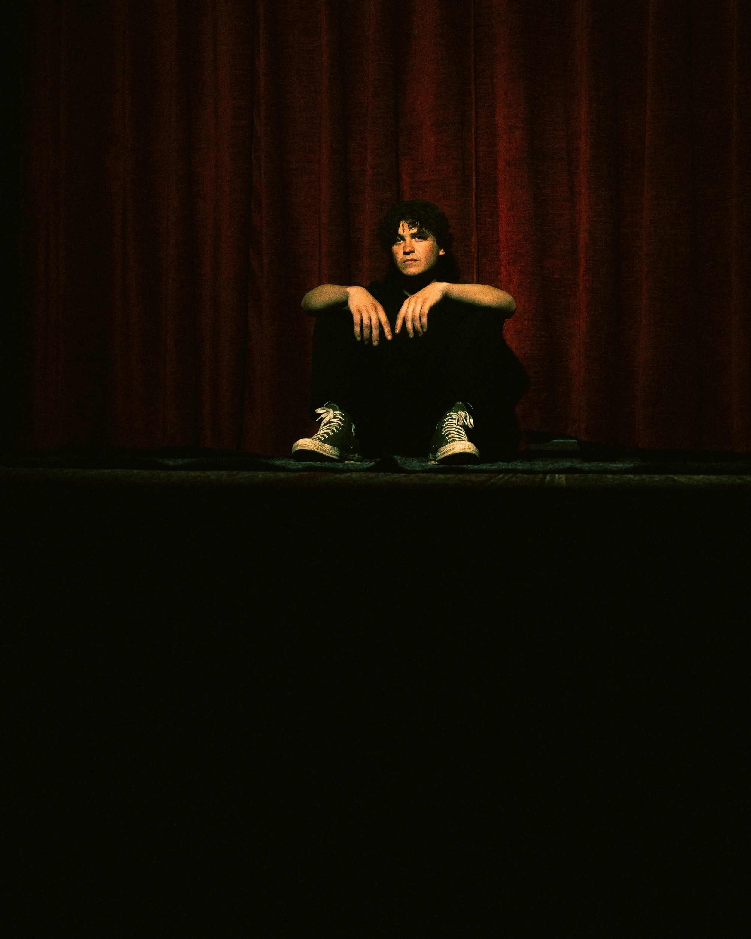 A person sitting on the floor with their knees up, arms resting on their knees, in front of a red curtain. They have curly dark hair and are wearing black clothing and sneakers.