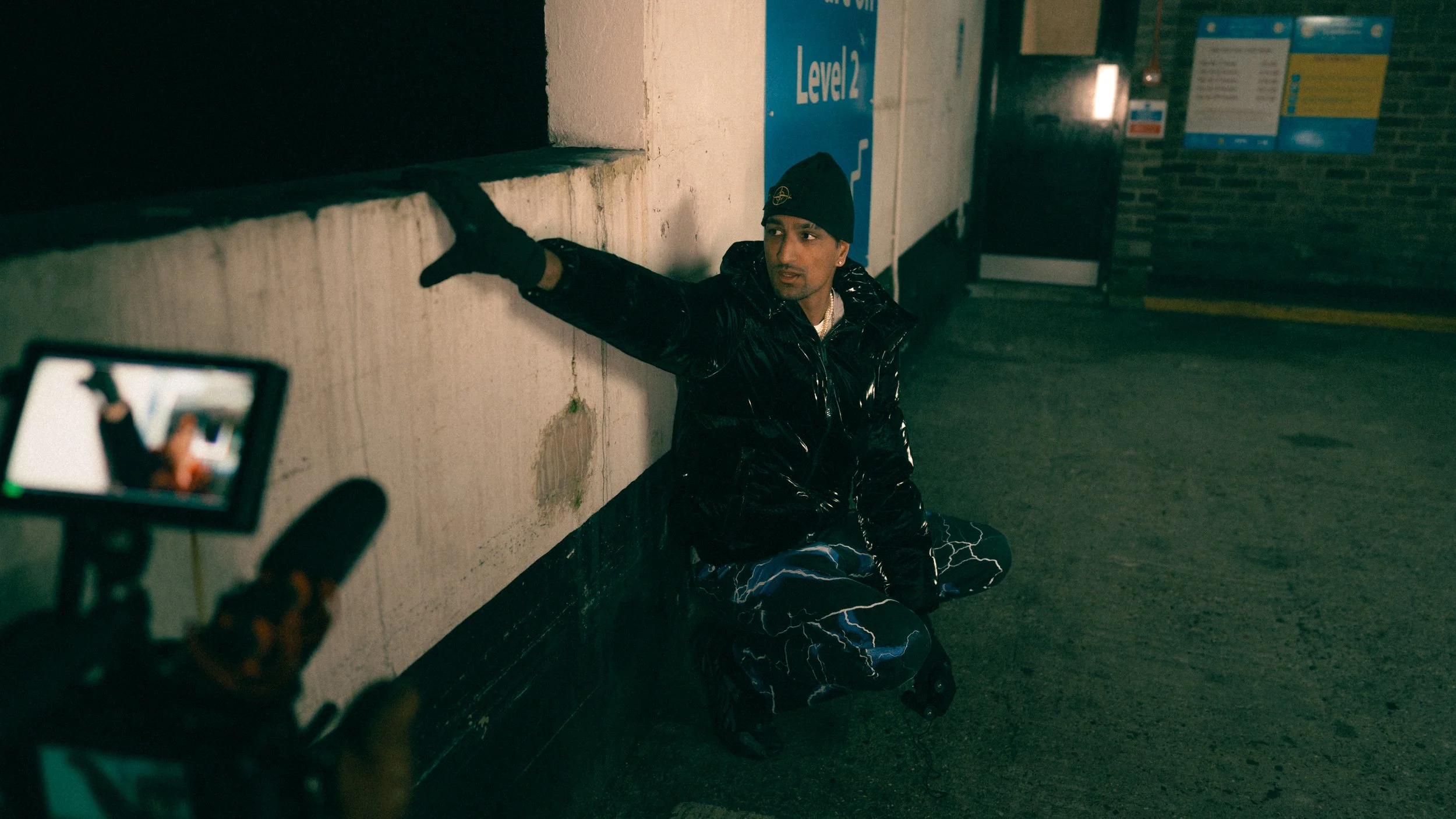 A young man wearing a black beanie, black shiny jacket, and lightning-patterned pants is sitting on the street leaning against a wall at night. A camera on a tripod is pointed at him.