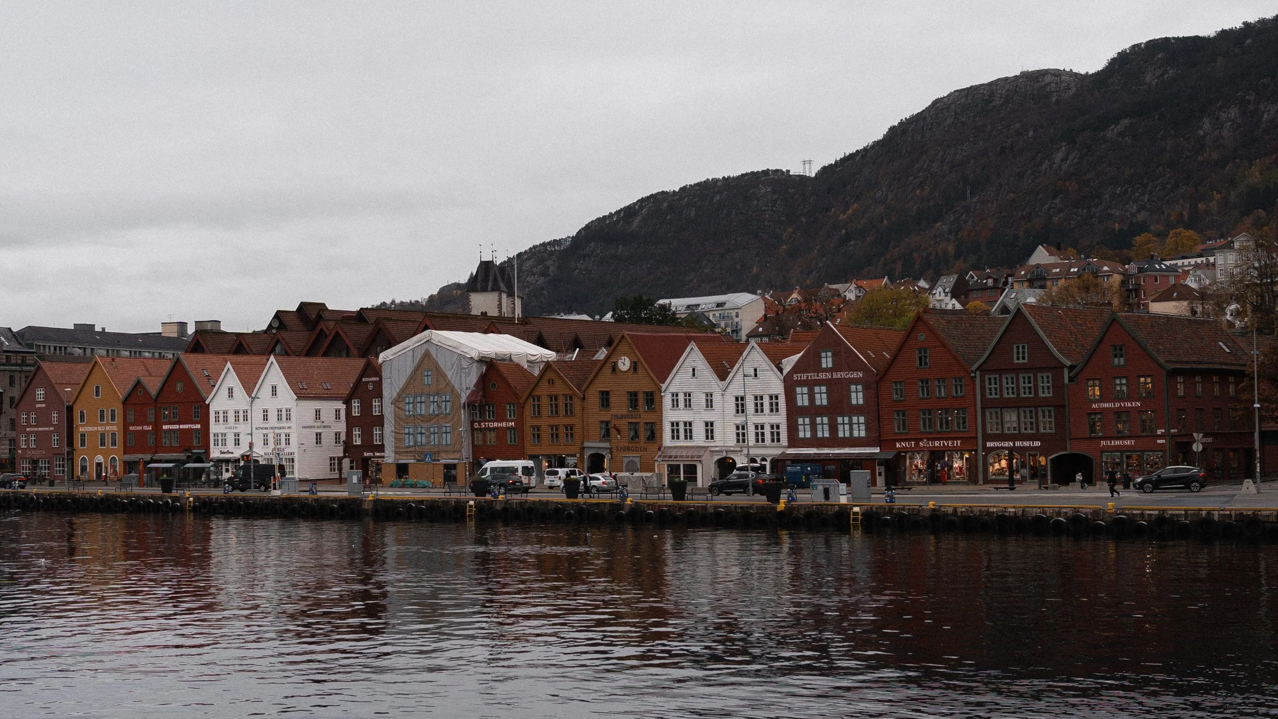 Colorful wooden buildings along a waterfront with mountains in the background on an overcast day.