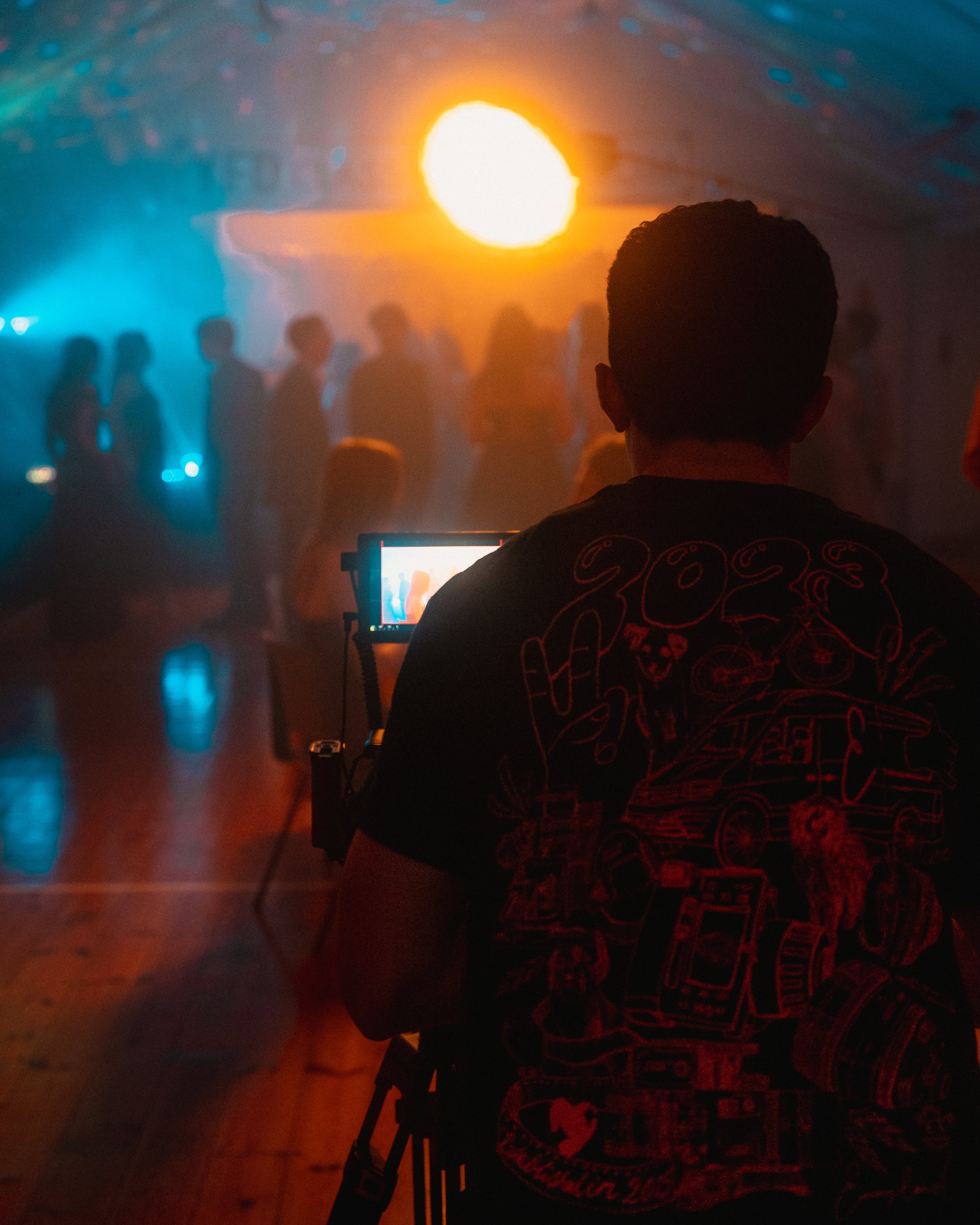 A person operating a camera in a dimly lit indoor space with people dancing or socializing in the background, illuminated by a large orange light or sunset glow.