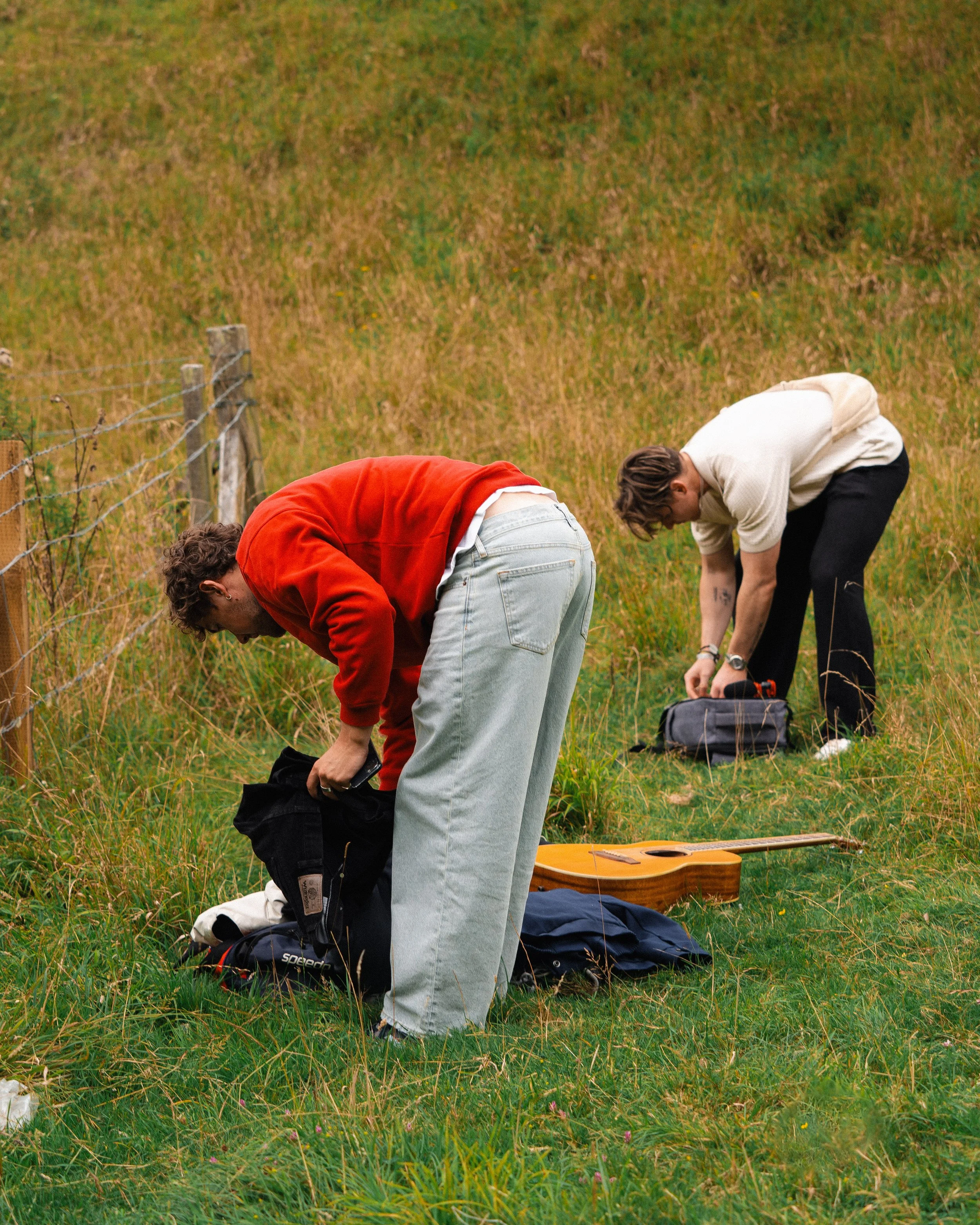 Two young men are kneeling on a grassy field, packing their backpacks with a guitar and outdoor gear nearby.