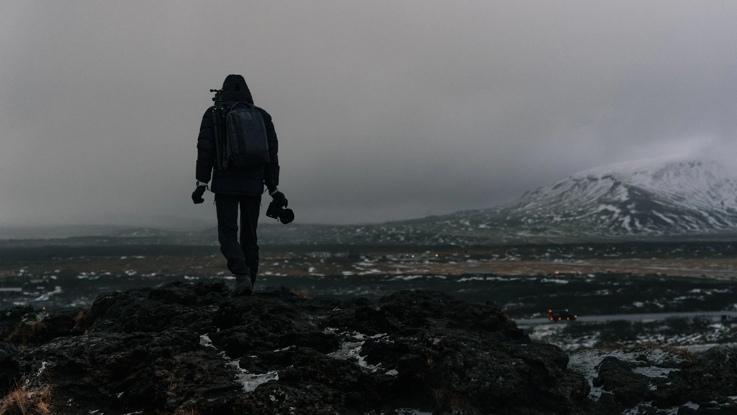 A person dressed in dark outdoor clothing, carrying a backpack and holding a helmet, walking on dark rocks in a cold, mountainous landscape with snow-covered peaks in the background under a cloudy sky.