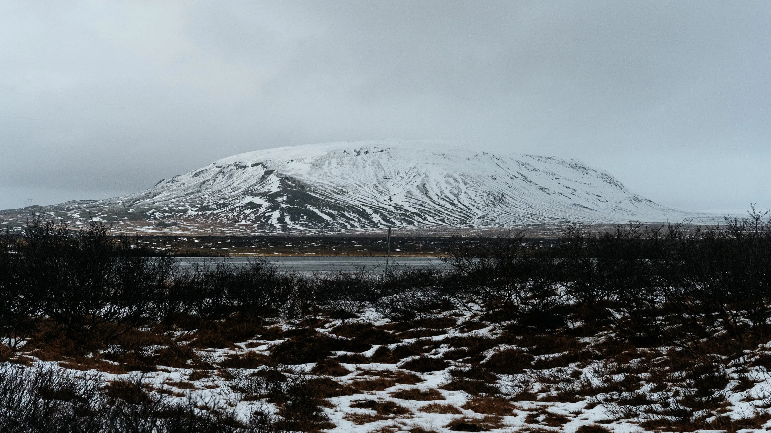 Snow-covered mountain with a dark, cloudy sky, sparse vegetation, and patches of snow on the ground.