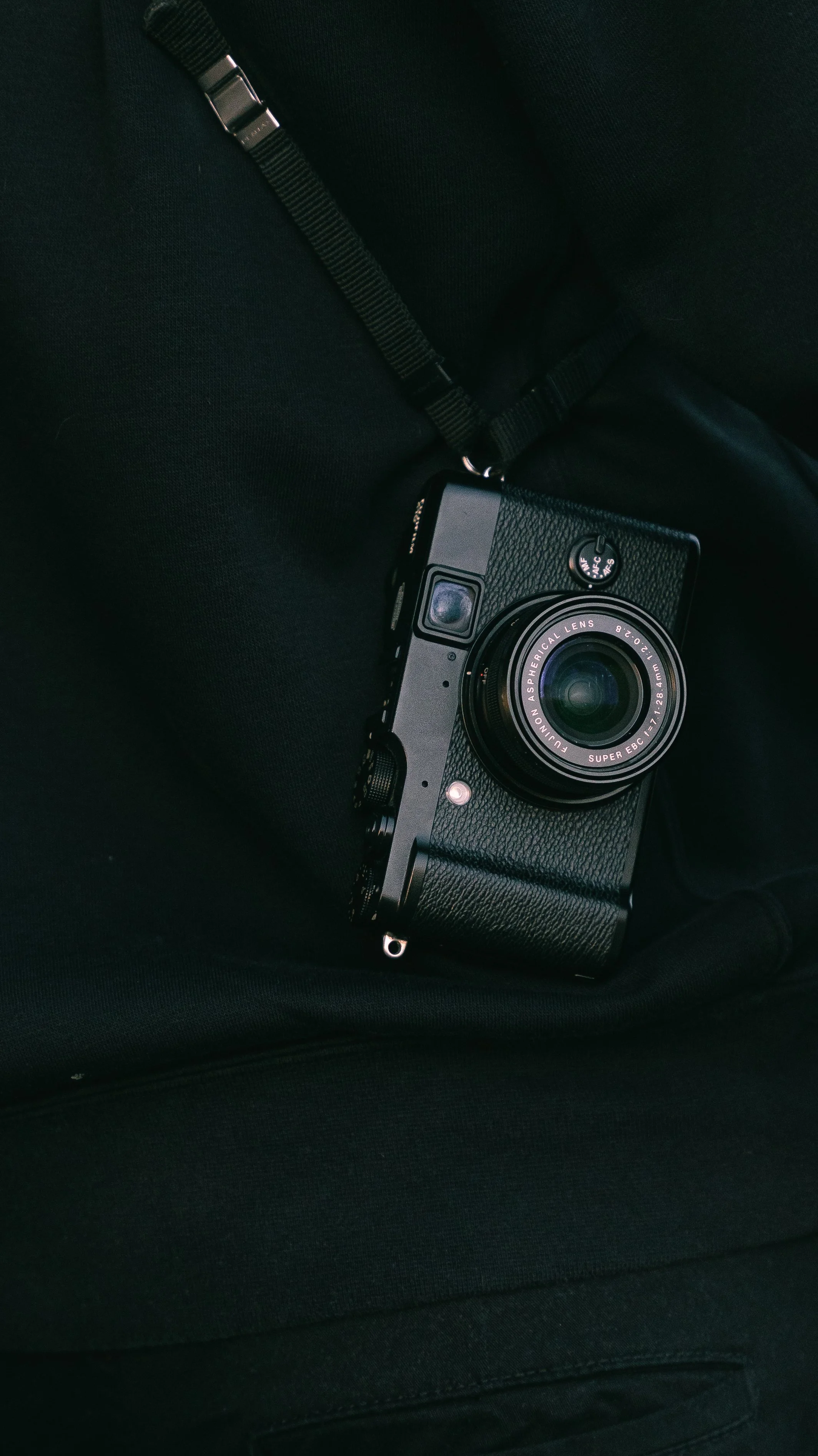A black compact camera with a lens, resting on a person's black clothing, with a camera strap attached.