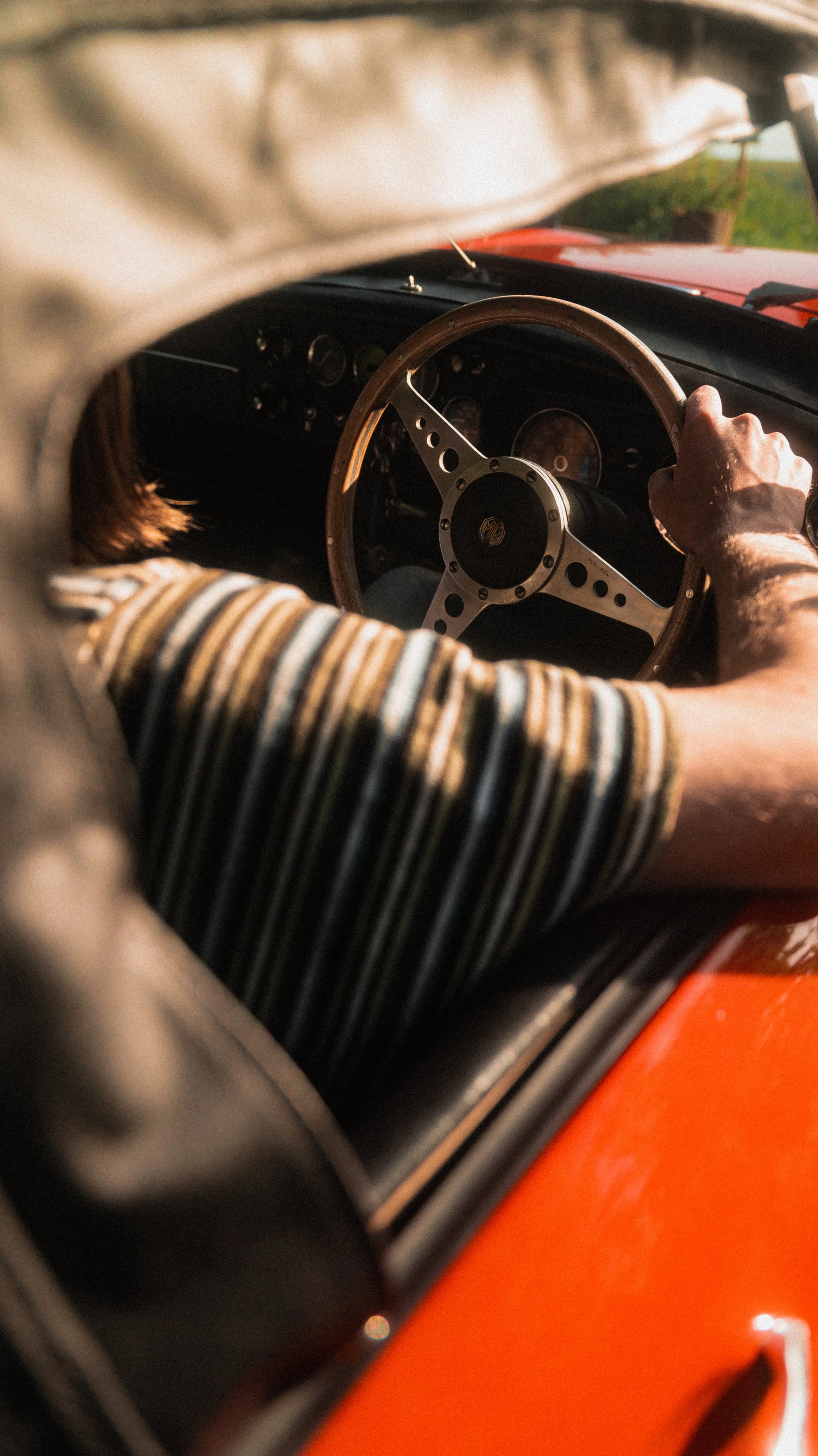 A person with a striped shirt driving a vintage red car, holding the wooden steering wheel, with a view of the dashboard through the windshield.
