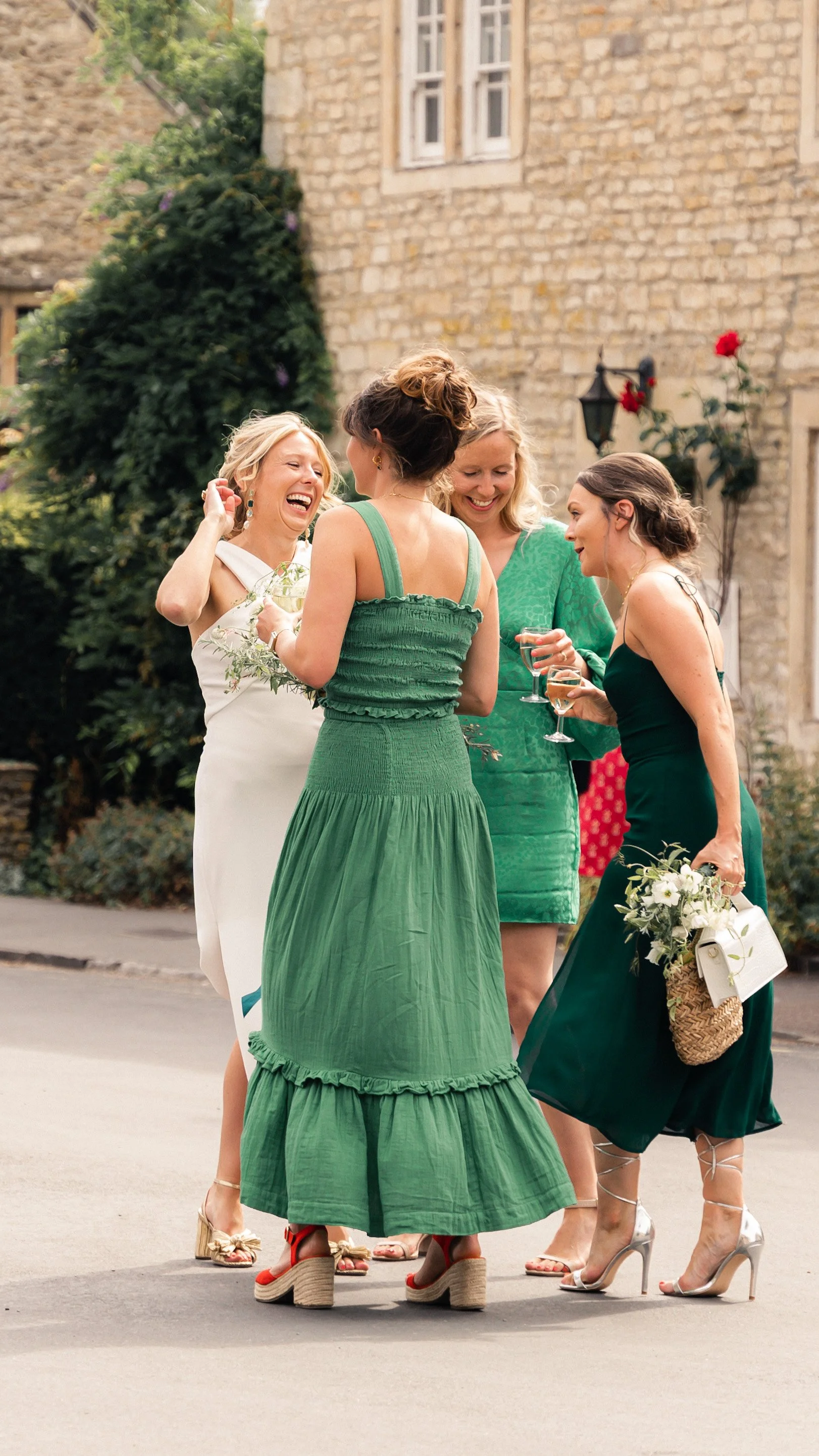 Four women dressed in elegant, colorful dresses are standing outside and chatting, with some holding glasses of wine. They are smiling and appear to be enjoying a social gathering, in front of a stone building with greenery and a lamp post.
