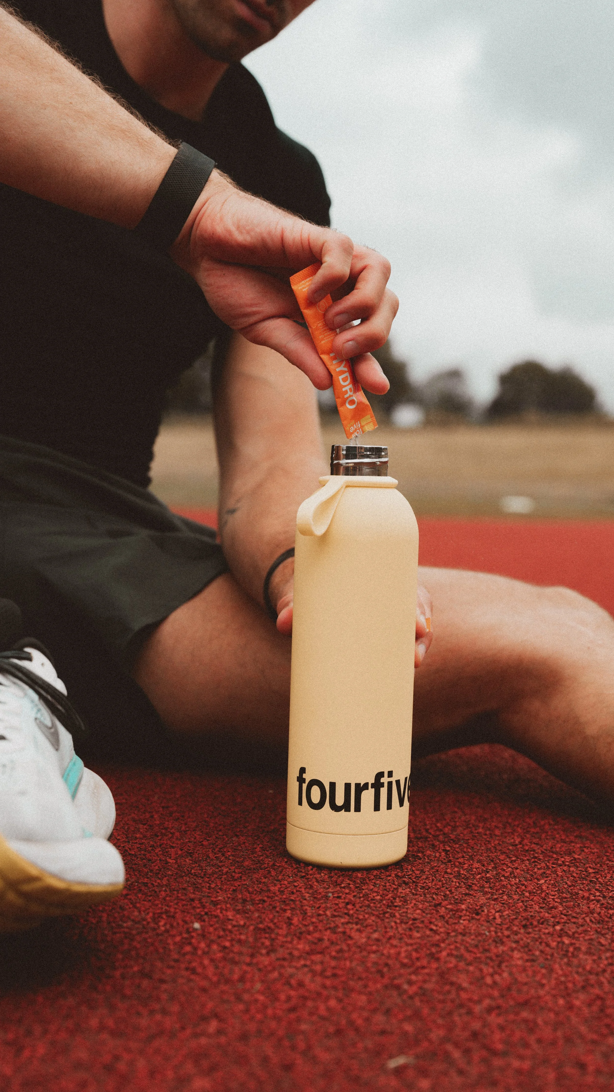 A person sitting outdoors on a red running track, opening a cream-colored water bottle with the brand name 'fourfives' printed at the bottom. The person's face is partially visible, and they are wearing casual athletic clothing and sneakers.