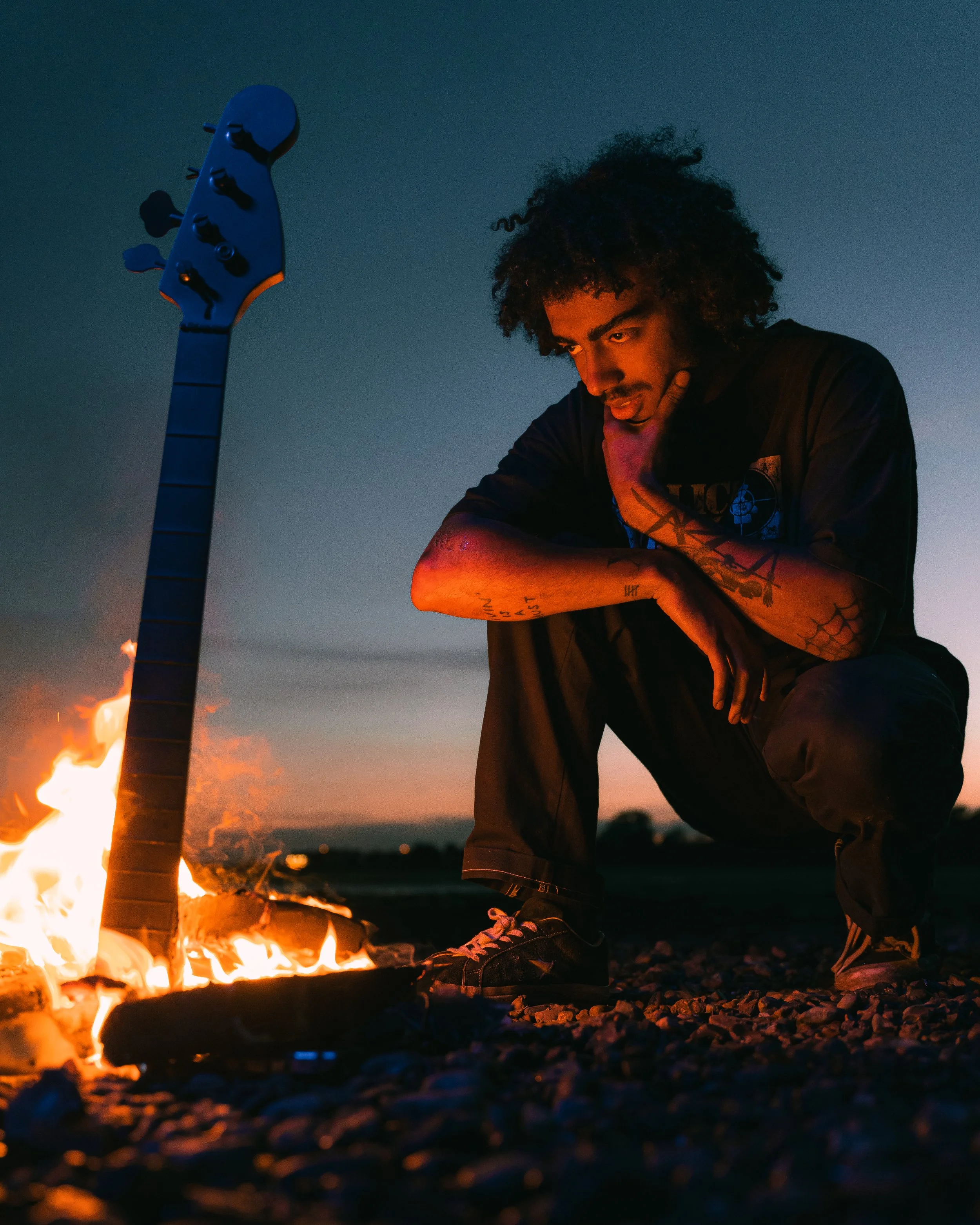 A man with tattoos crouching near a campfire at dusk with a guitar standing upright next to him.