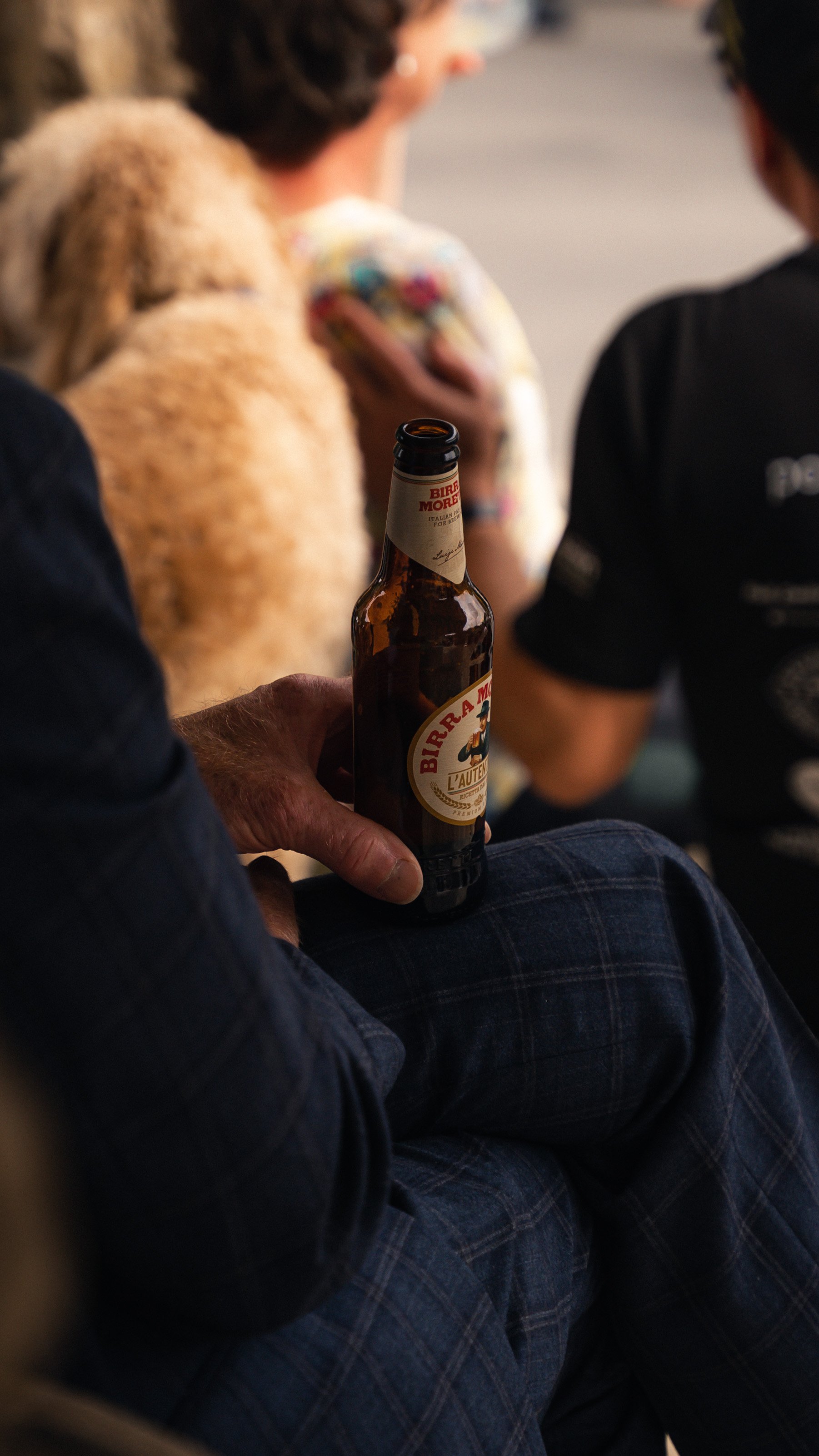 A person holding an open bottle of Birra Moretti beer, sitting with crossed legs in a crowd, with women in the background.